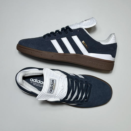 A pair of Adidas Skateboarding Busenitz sneakers in navy blue suede with white stripes, white laces, a white tongue, and a brown gum sole, displayed on a light gray background.