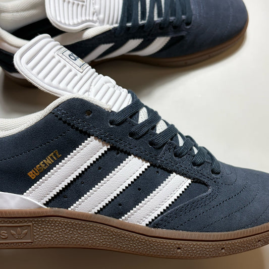 A close-up of an Adidas Busenitz  sneaker in navy blue suede with white stripes, a white tongue, and a brown gum sole. The Adidas logo is visible on the side heel, and 'BUSSNITZ' is printed in gold on the side.