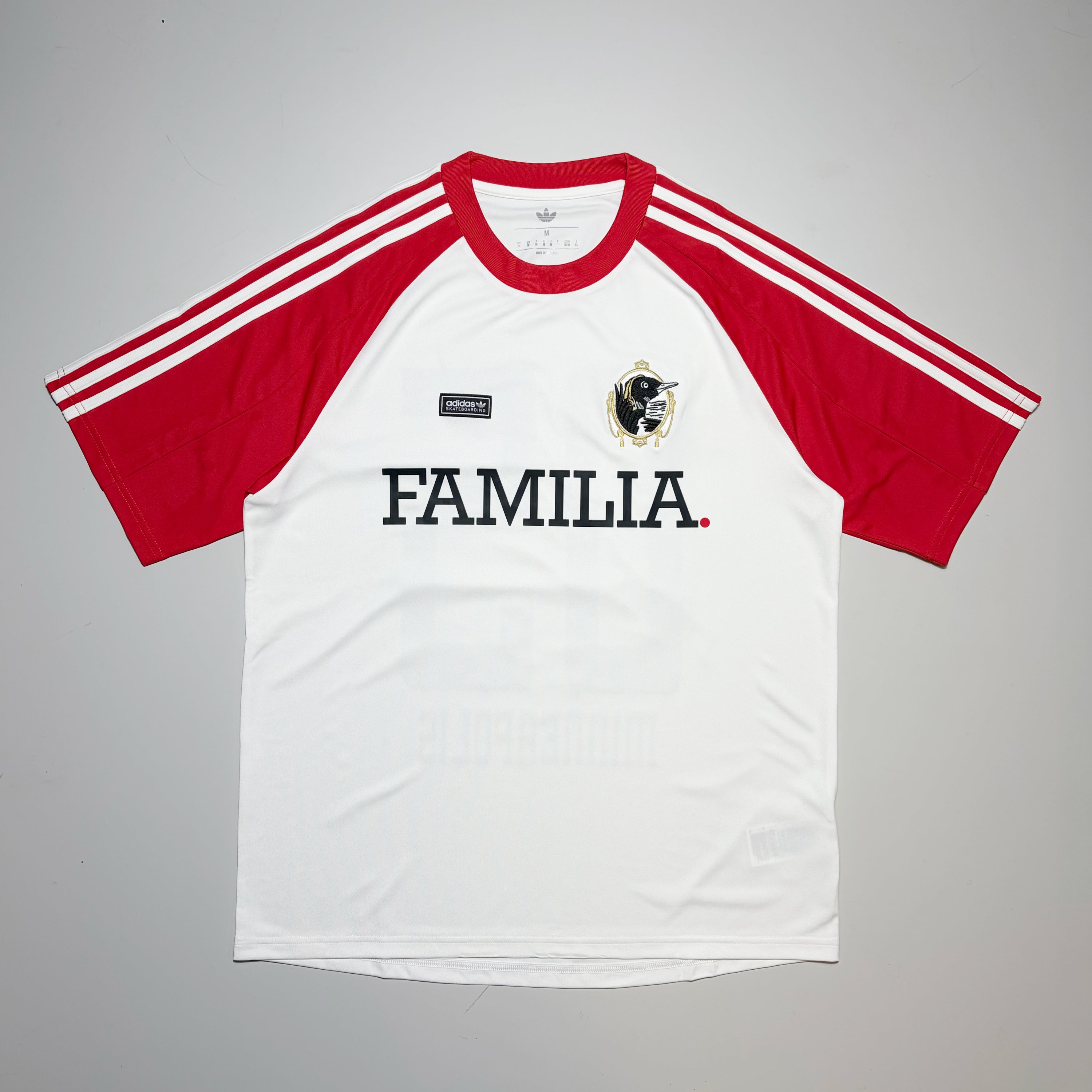 A white and red short-sleeve raglan soccer jersey with 'FAMILIA' printed across the chest, featuring three white stripes on the sleeves and a black-and-white loon crest on the left chest.