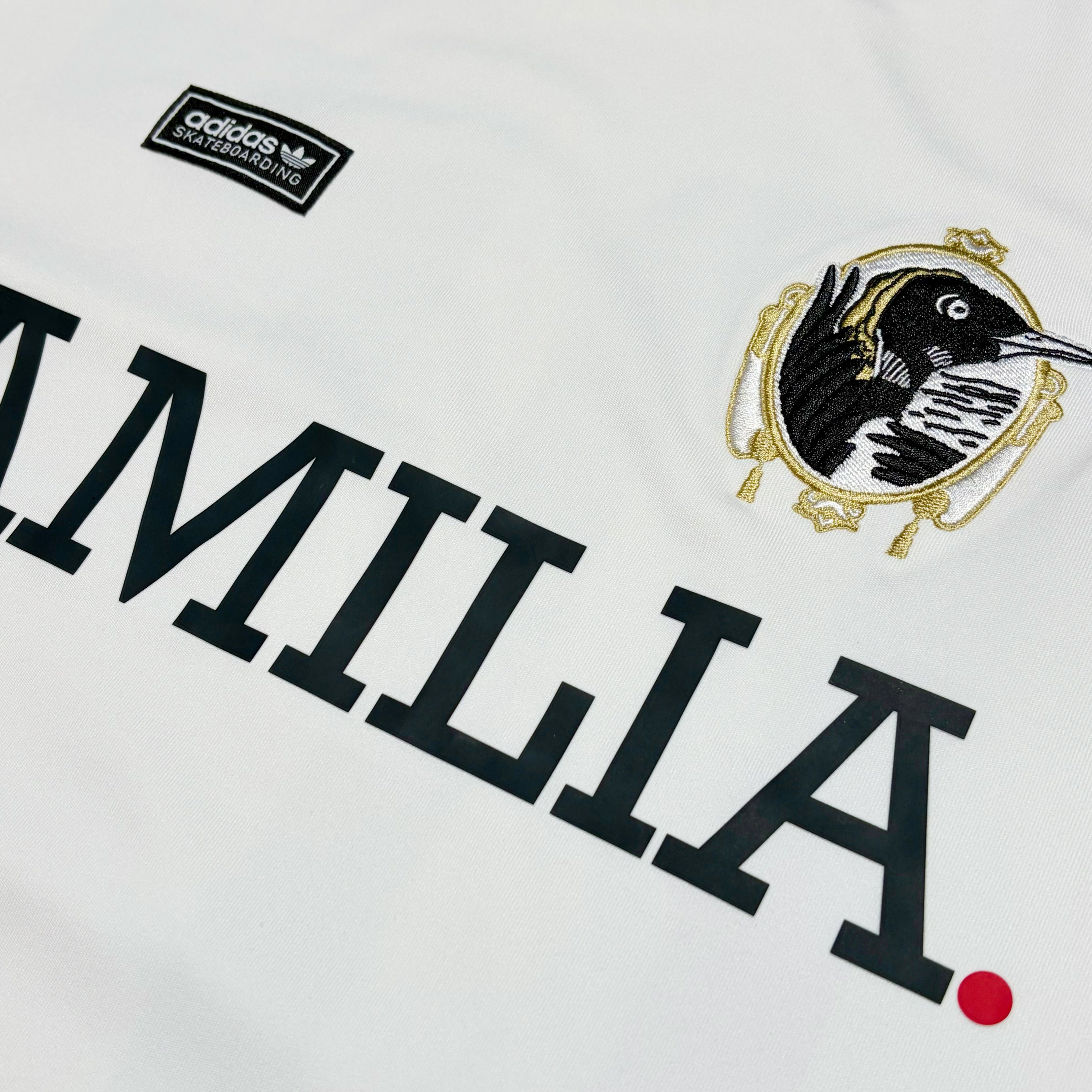 Close-up of a white Adidas soccer jersey featuring the word 'FAMILIA' in bold black lettering, an Adidas logo patch, and a black bird crest with gold detailing.