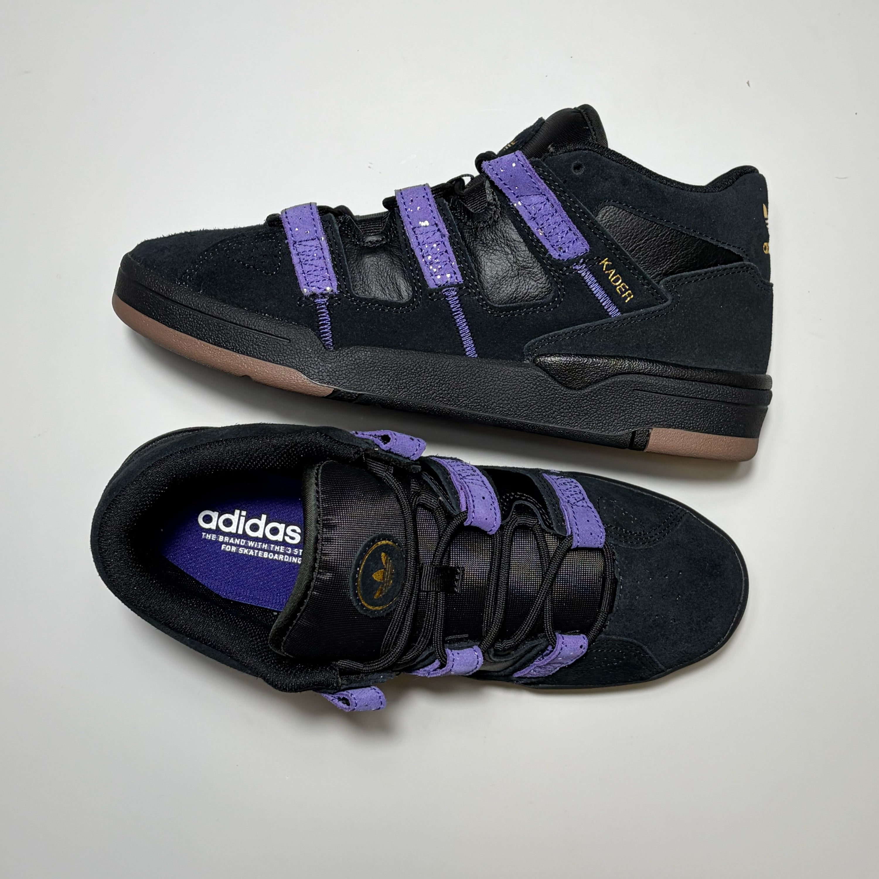 A pair of Adidas Skateboarding Kader Sylla Pro model sneakers in black suede and leather with purple straps and a tan outsole, displayed on a light gray background.