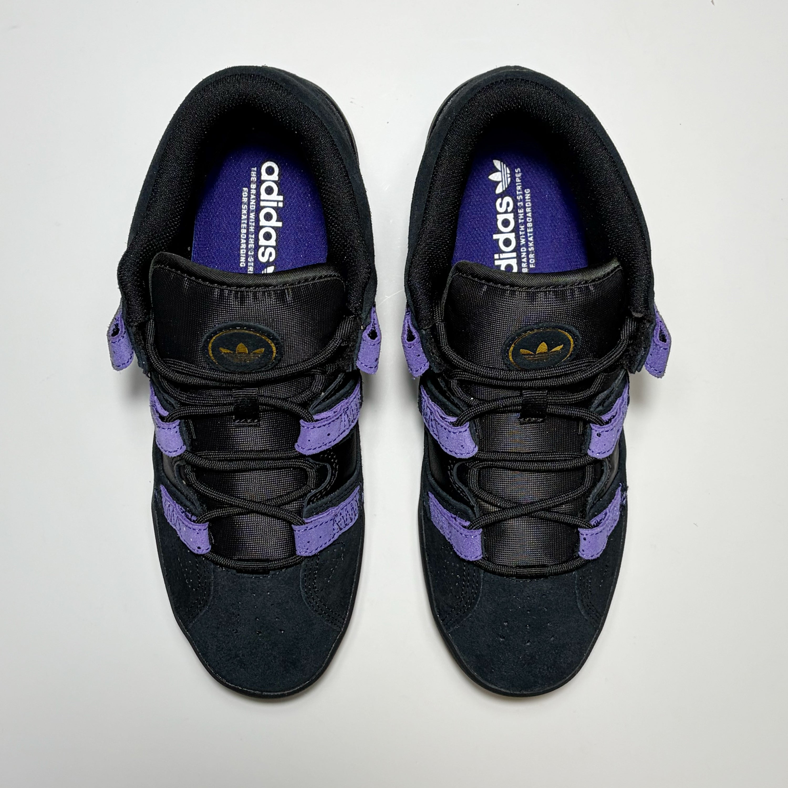 A pair of Adidas Kader sneakers shown from a top-down view. The shoes are primarily black with prominent purple accents on the laces, side straps, and inner lining, and a gold logo on the tongue.