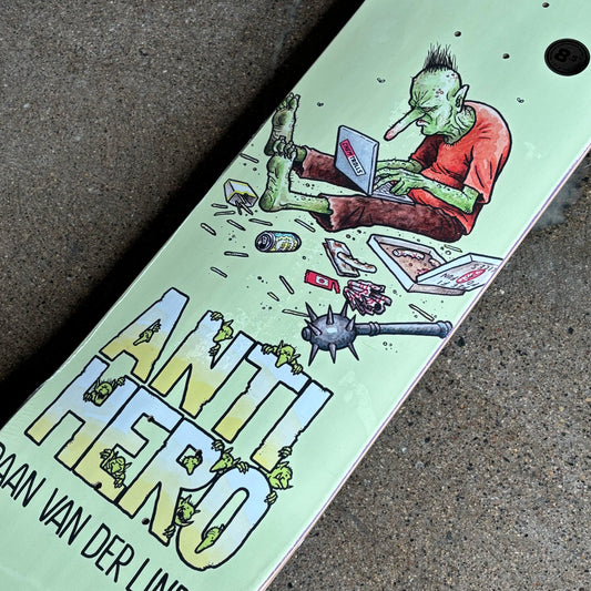 Skateboard deck with a graphic of a green-skinned person using a laptop, surrounded by objects like a bottle and a weapon, on a 'Anti Hero' branded deck.