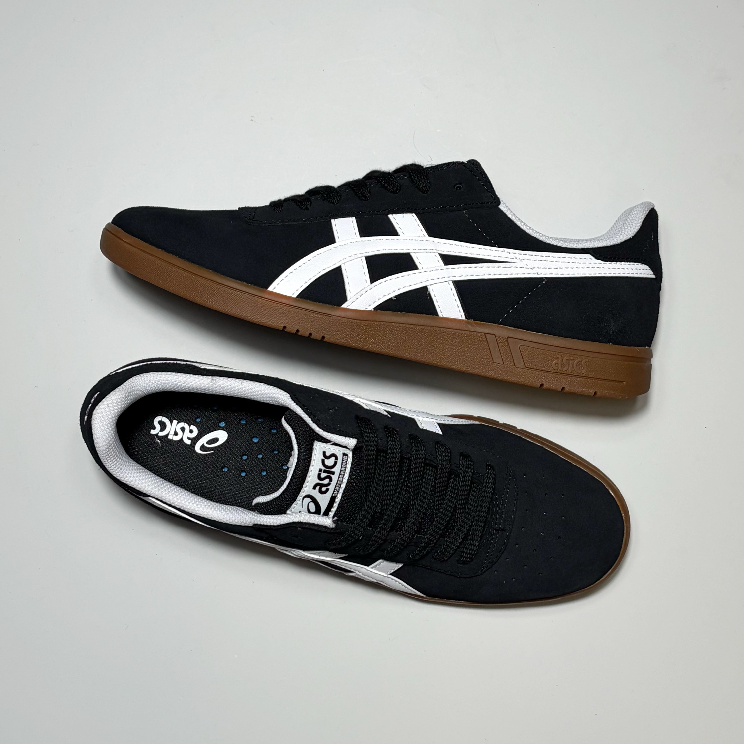 A pair of Asics skate shoes in black suede with white stripes and brown gum soles, displayed on a light gray background.
