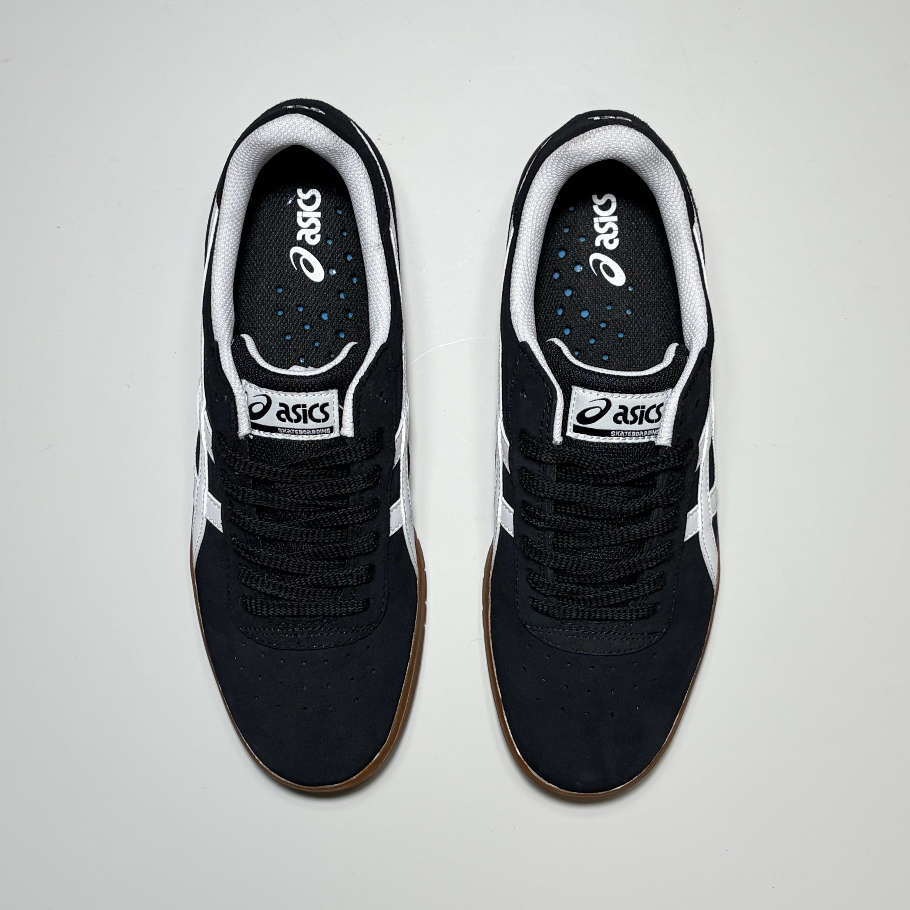 A pair of ASICS sneakers shown from a top-down view on a light gray background. The shoes are primarily black with white side stripes, white tongue labels, and a brown gum outsole.