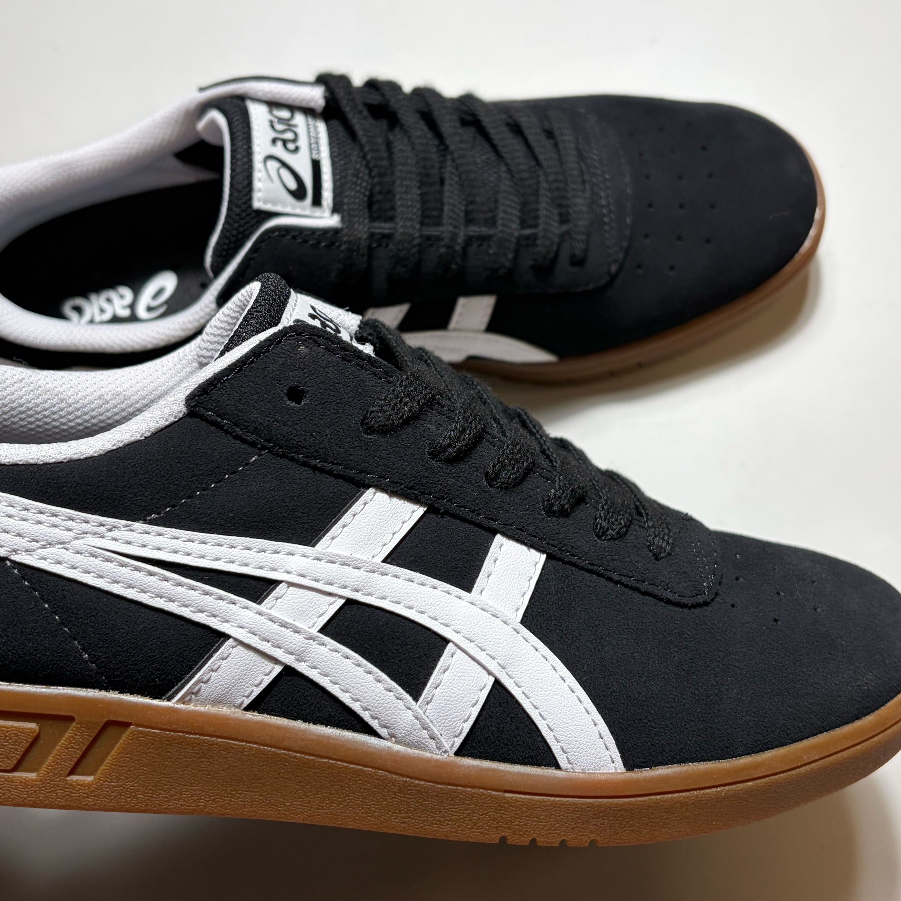 Black and white Asics skate shoes with a brown gum sole, shown on a light off-white background.