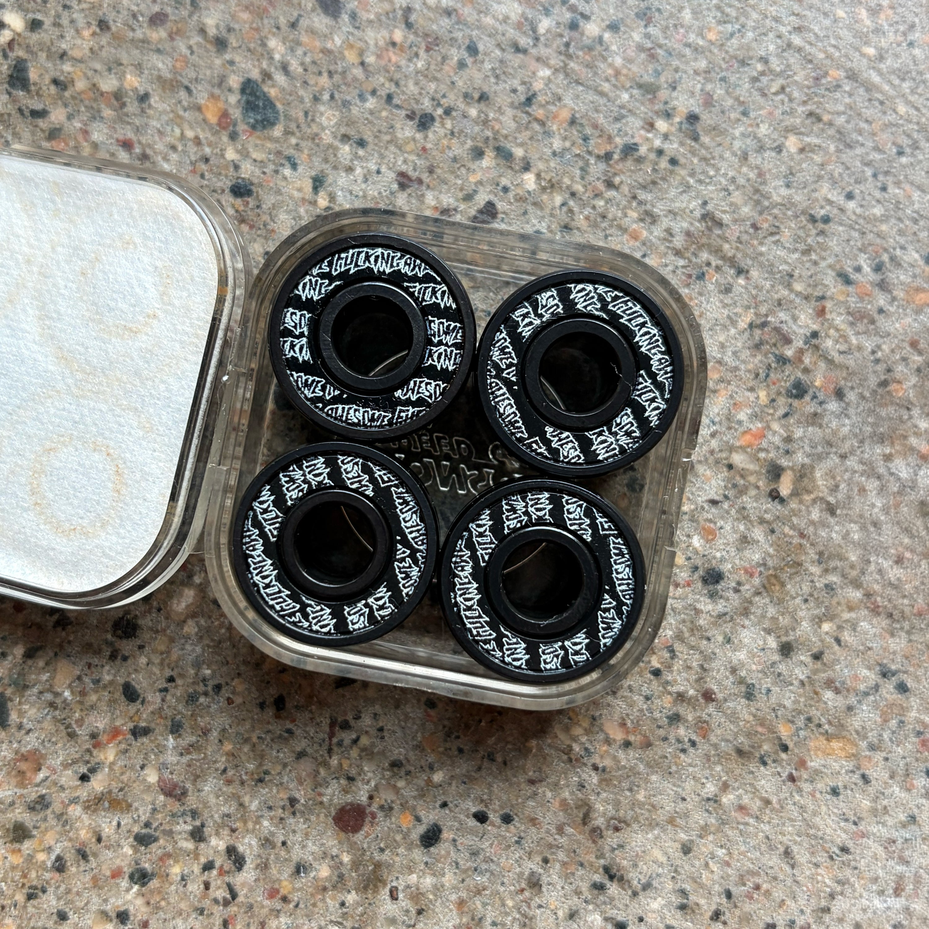 Four black skateboard bearings with white script logos, displayed in a clear plastic case on a speckled granite countertop.