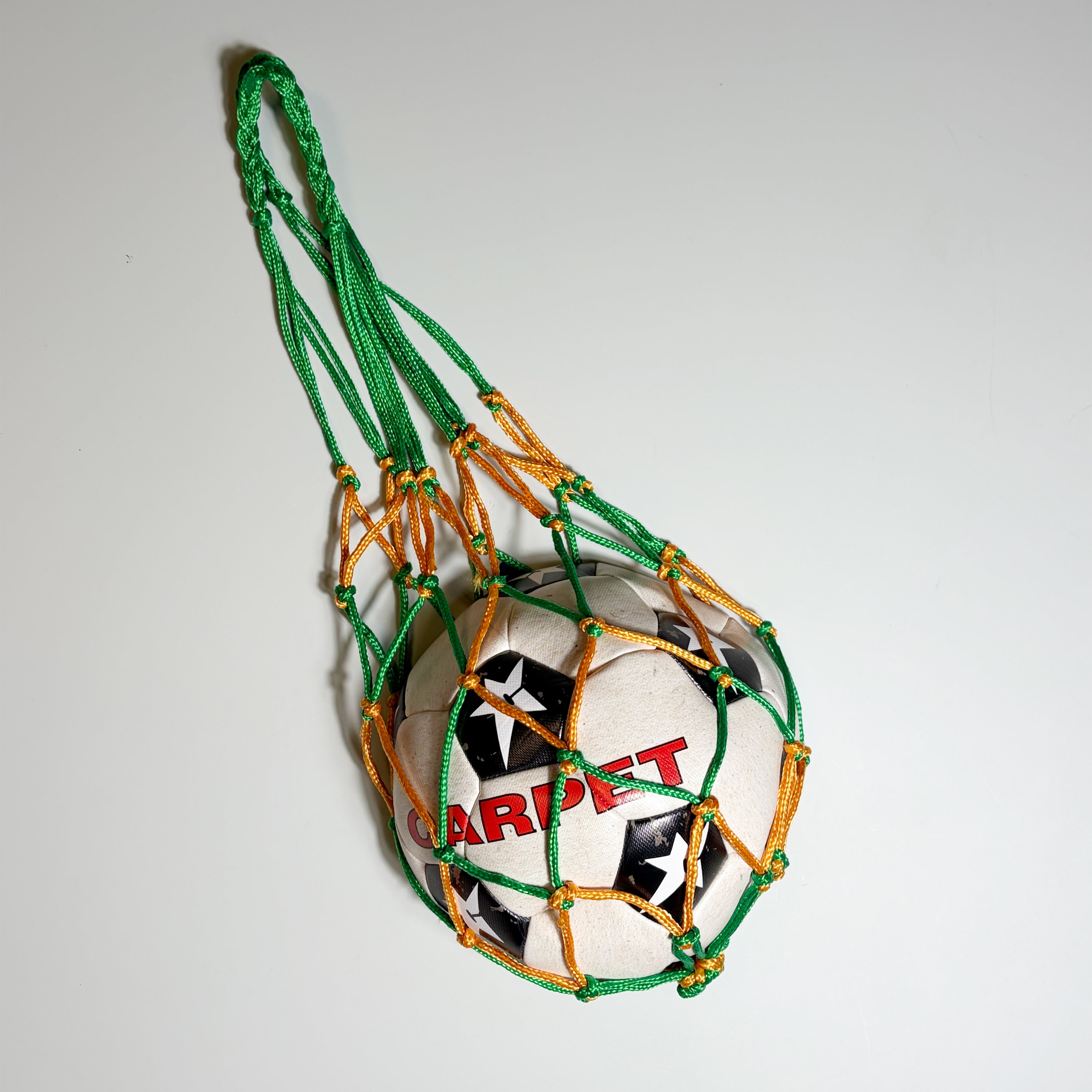 A soccer ball with 'CARPET' printed in red on its surface, suspended inside a green and orange net with a long green loop at the top, photographed against a plain light background.