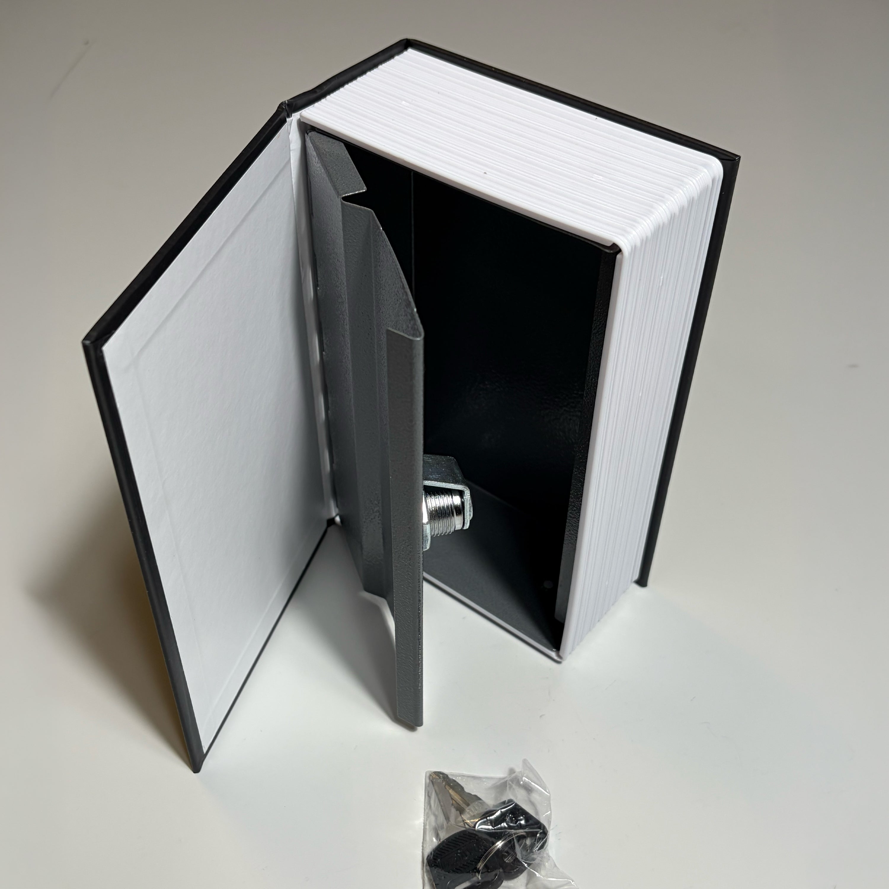 A book-shaped safe with a white striped cover and black spine, shown open to reveal a dark interior and a metal lock mechanism. A small plastic bag with keys is placed on the surface below.