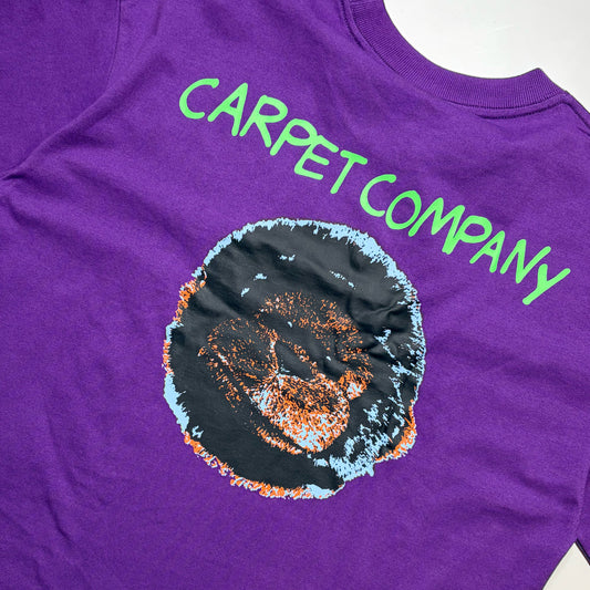 A close-up of a purple t-shirt featuring a graphic of a black circular carpet with orange and light blue speckles, and the words 'CARPET COMPANY' printed in bright green above it.