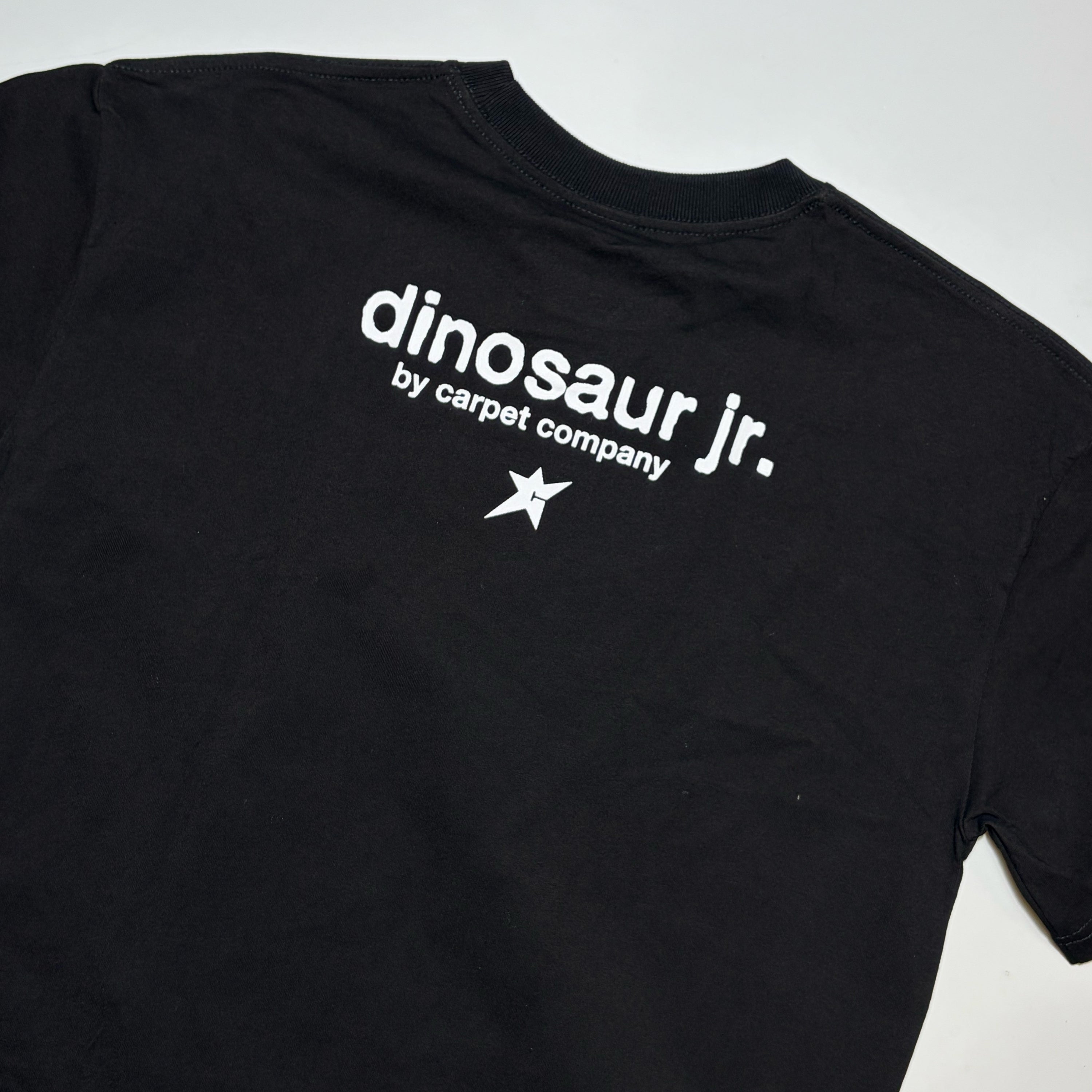 A black short-sleeve t-shirt laid flat on a light gray background, featuring the white printed text 'dinosaur jr. by carpet company' and a small white star logo on the chest.