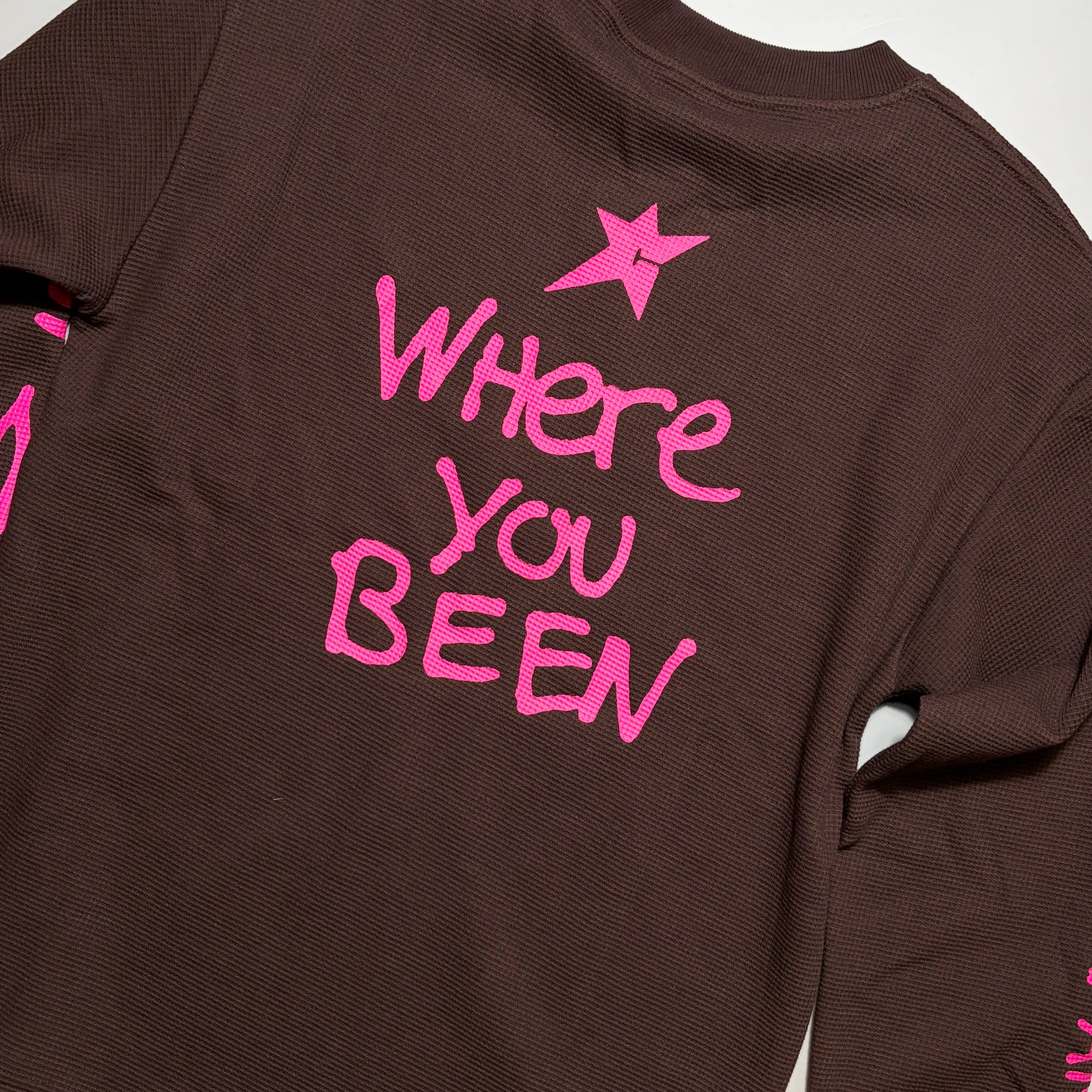 A close-up of a dark brown ribbed long-sleeve shirt showing the back, decorated with a bright pink star and the text 'Where You Been' in matching pink lettering.