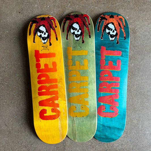 Three skateboard deck tops displayed vertically on a concrete floor, each featuring a red spider and skull graphic above the word 'CARPET' in large vertical lettering. The left deck is yellow with red lettering, the middle is olive green with orange lettering, and the right is teal with pink lettering.