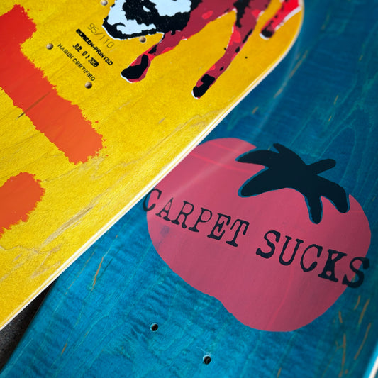 Two skateboard decks shown diagonally, one with a yellow background and orange graphic, and the other with a blue wood grain background and a pink tomato graphic containing the text 'CARPET SUCKS'.