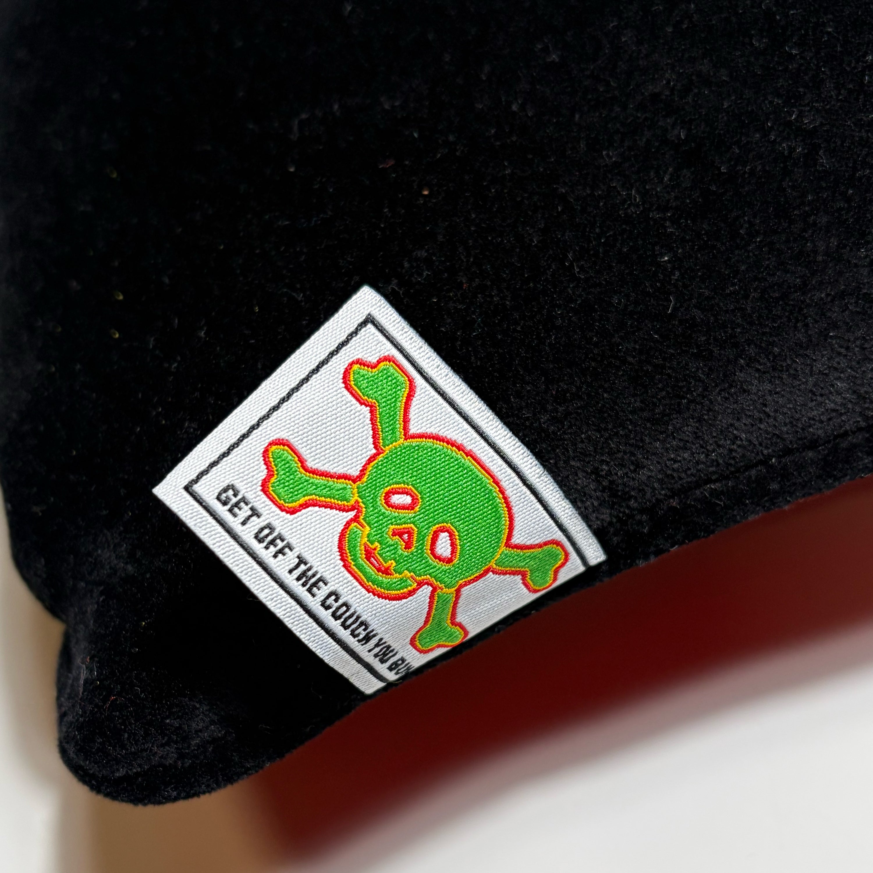 Close-up of a black plush or velvety fabric item with a sewn square label showing a green skull-and-crossbones graphic outlined in red and the text 'GET OFF THE COUCH NOW'.
