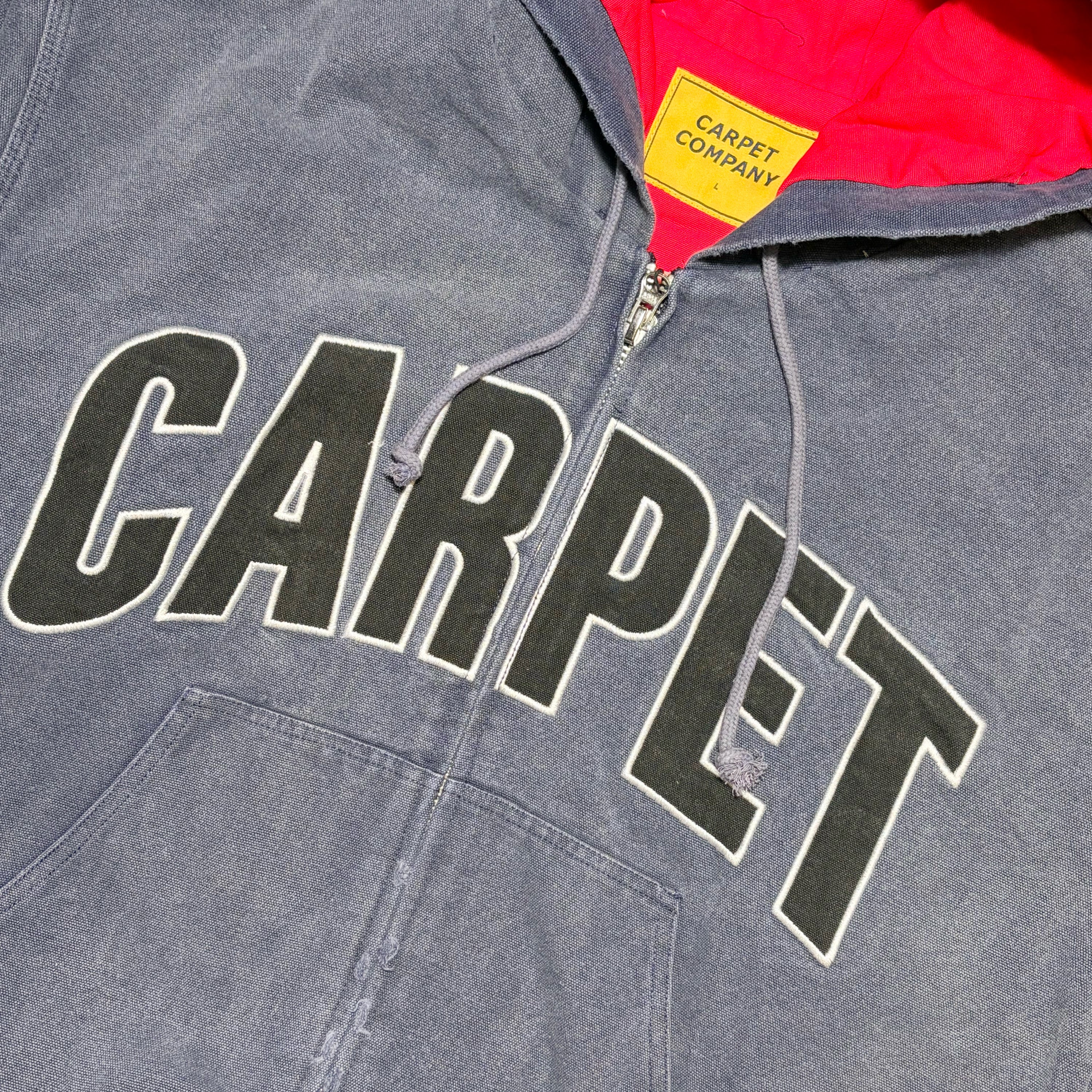 Close-up of a gray hooded sweatshirt with large black 'CARPET' lettering outlined in white across the chest, a red hood lining, and a yellow neck label reading 'CARPET COMPANY'.