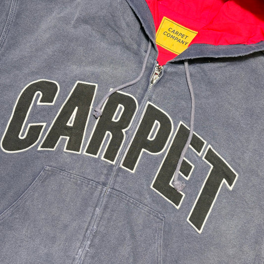 Close-up of a gray hooded sweatshirt with large black 'CARPET' lettering outlined in white across the chest, a red hood lining, and a yellow neck label reading 'CARPET COMPANY'.