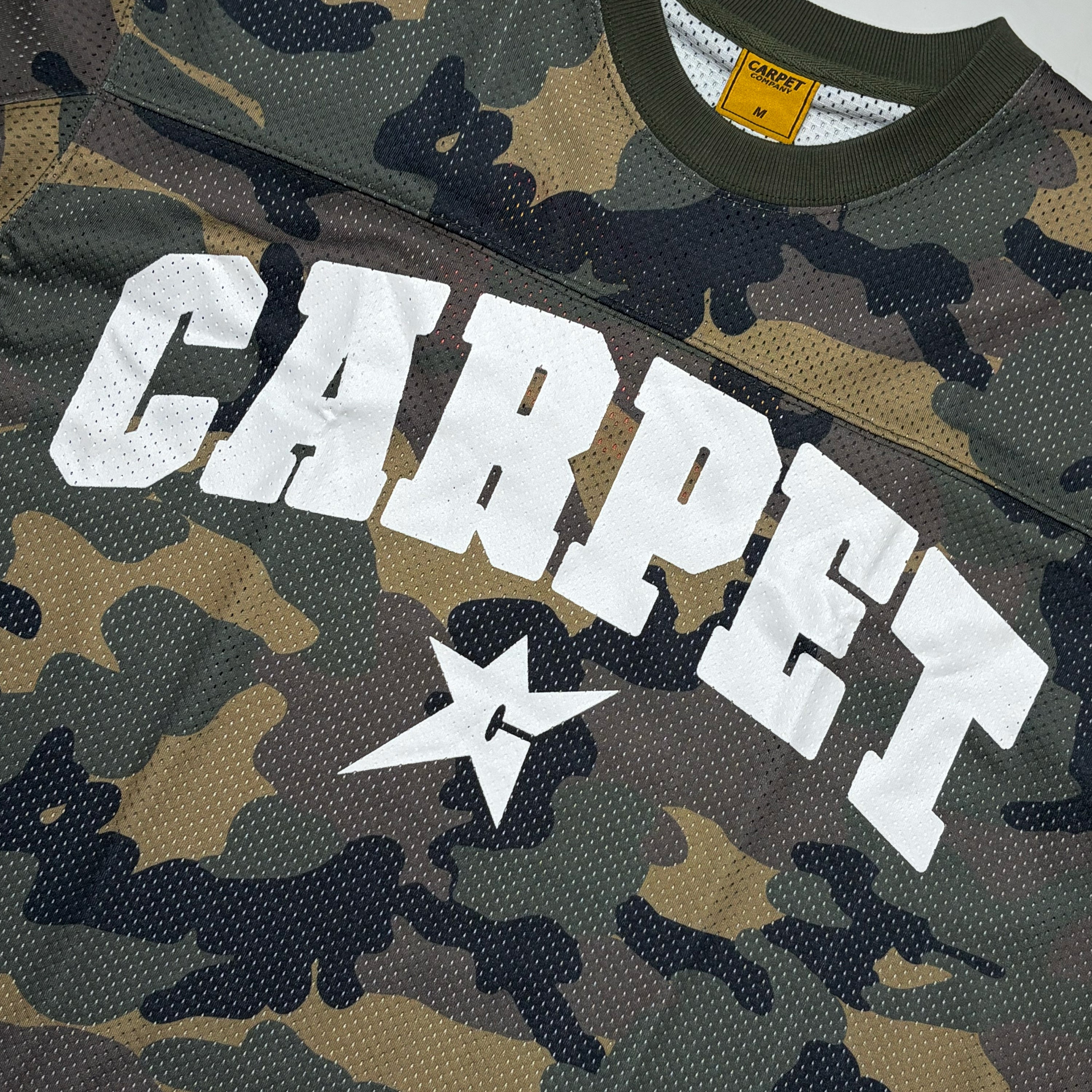 Close-up of a camouflage mesh jersey with large white 'CARPET' lettering and a white star logo on the front. The shirt has a green collar and a yellow neck label.