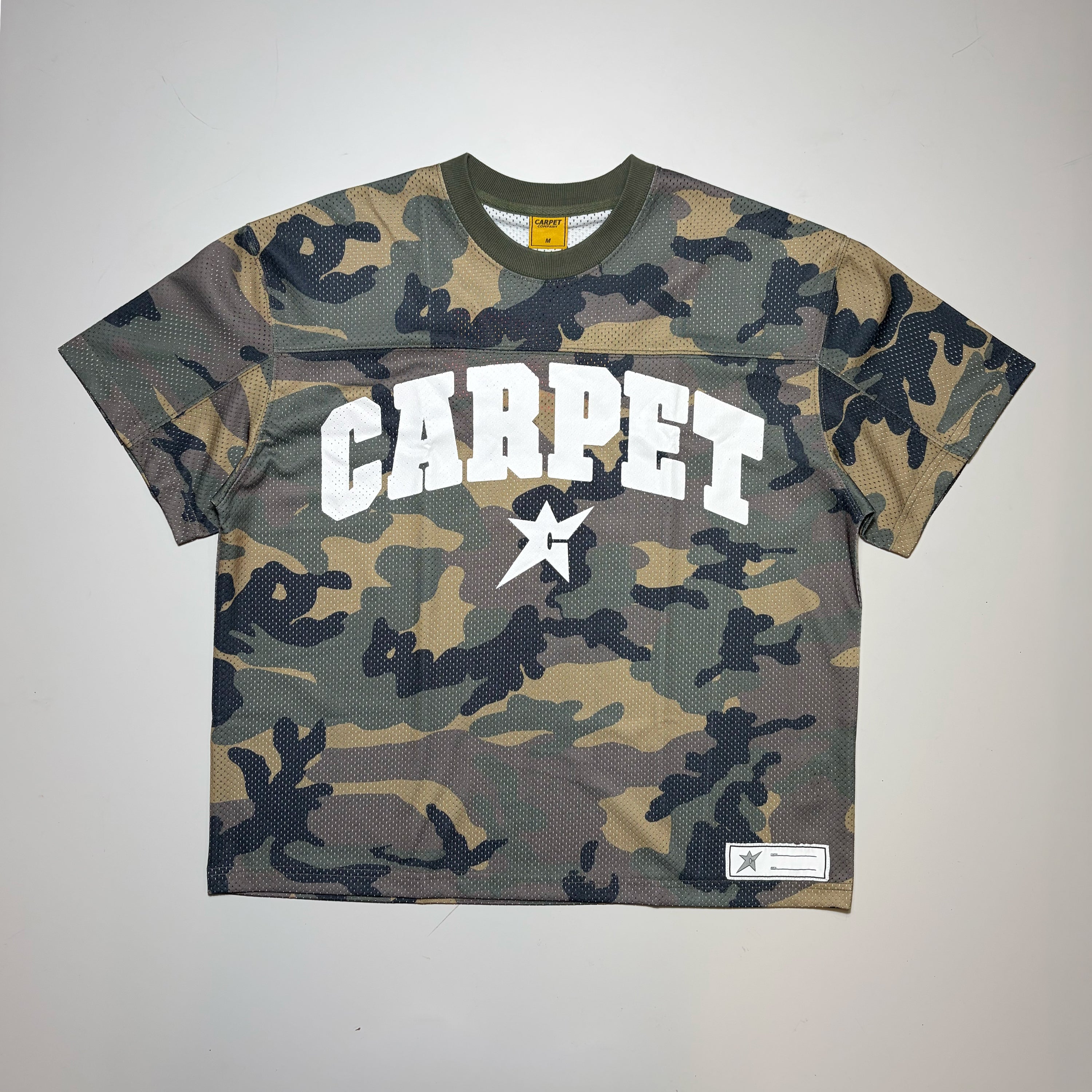 A short-sleeve cropped Carpet Company Jersey with a green, brown, and black camouflage pattern. The front features the word 'CARPET' in bold white varsity letters with a white star graphic in the center, and a small white brand patch on the bottom right hem.