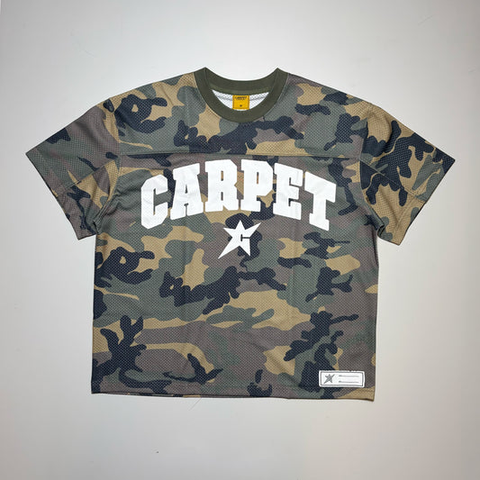 A short-sleeve cropped Carpet Company Jersey with a green, brown, and black camouflage pattern. The front features the word 'CARPET' in bold white varsity letters with a white star graphic in the center, and a small white brand patch on the bottom right hem.