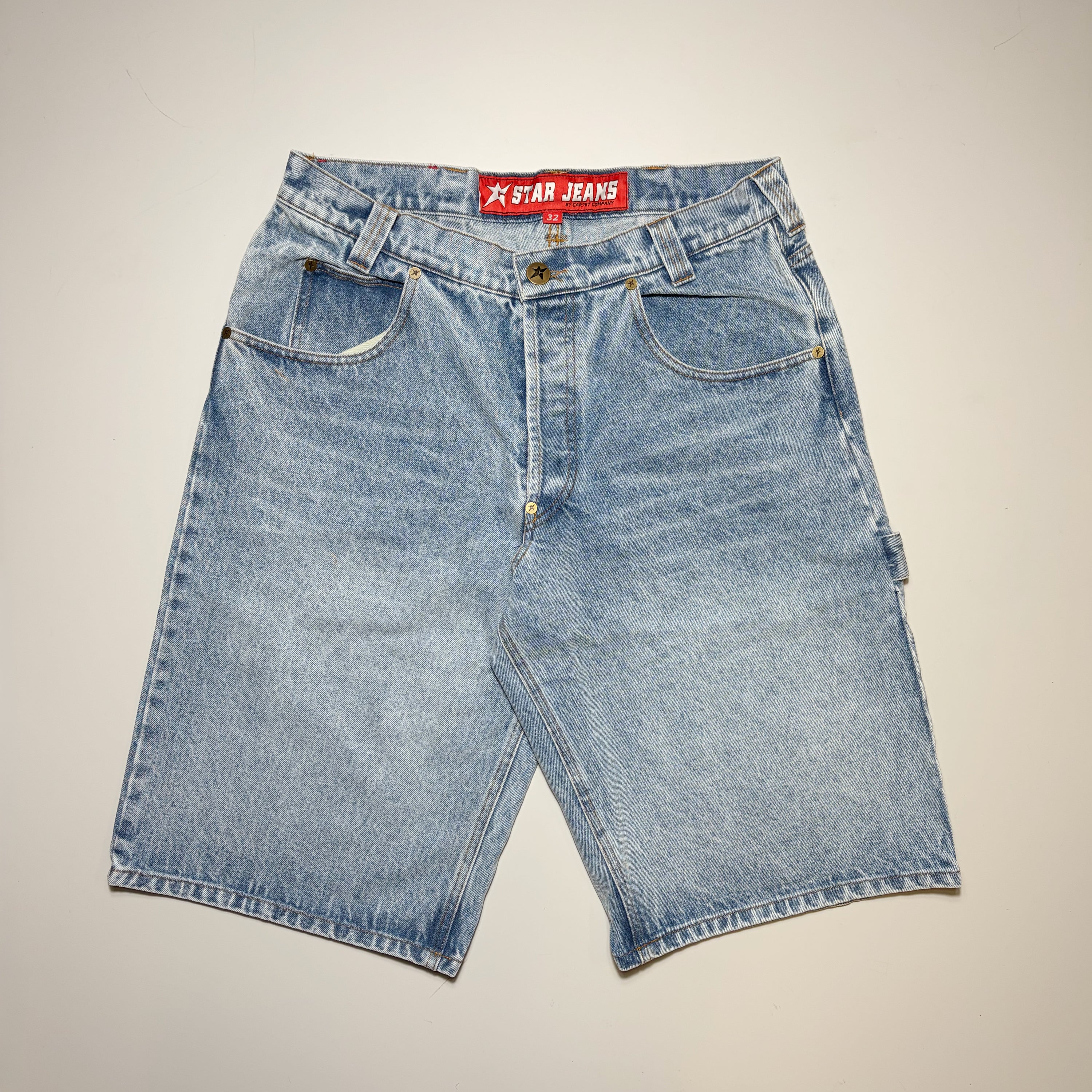 Light blue denim shorts with a faded wash, featuring a red waistband label that reads 'STAR JEANS' in white text, laid flat against a light beige background.