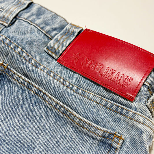 Close-up of the back waistband of light-wash blue denim jeans with a bright red leather patch embossed with the brand name 'STAR JEANS'.