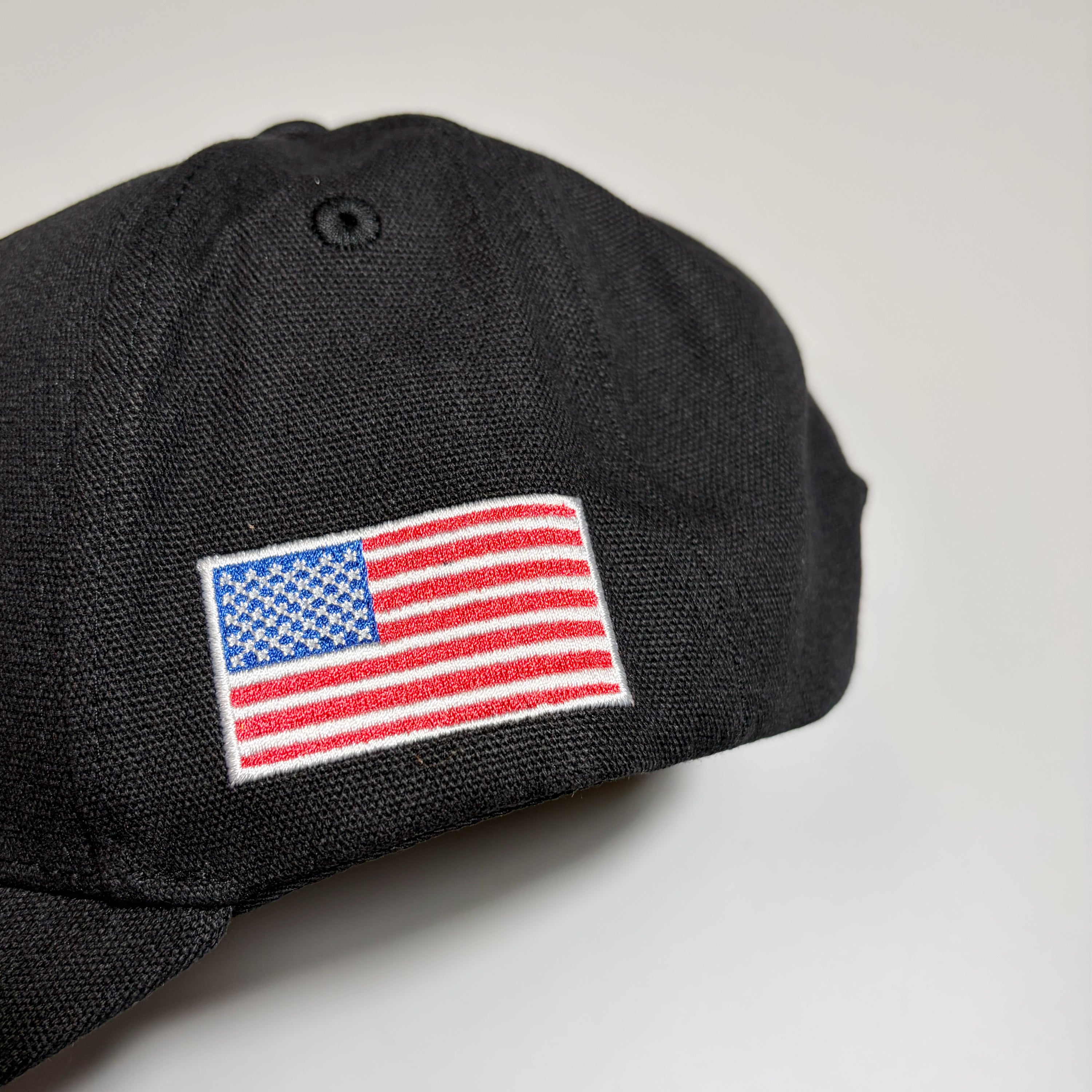 A close-up of a black baseball cap featuring an embroidered American flag patch on the side, set against a plain light gray background.
