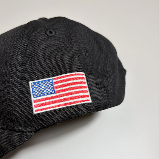A close-up of a black baseball cap featuring an embroidered American flag patch on the side, set against a plain light gray background.