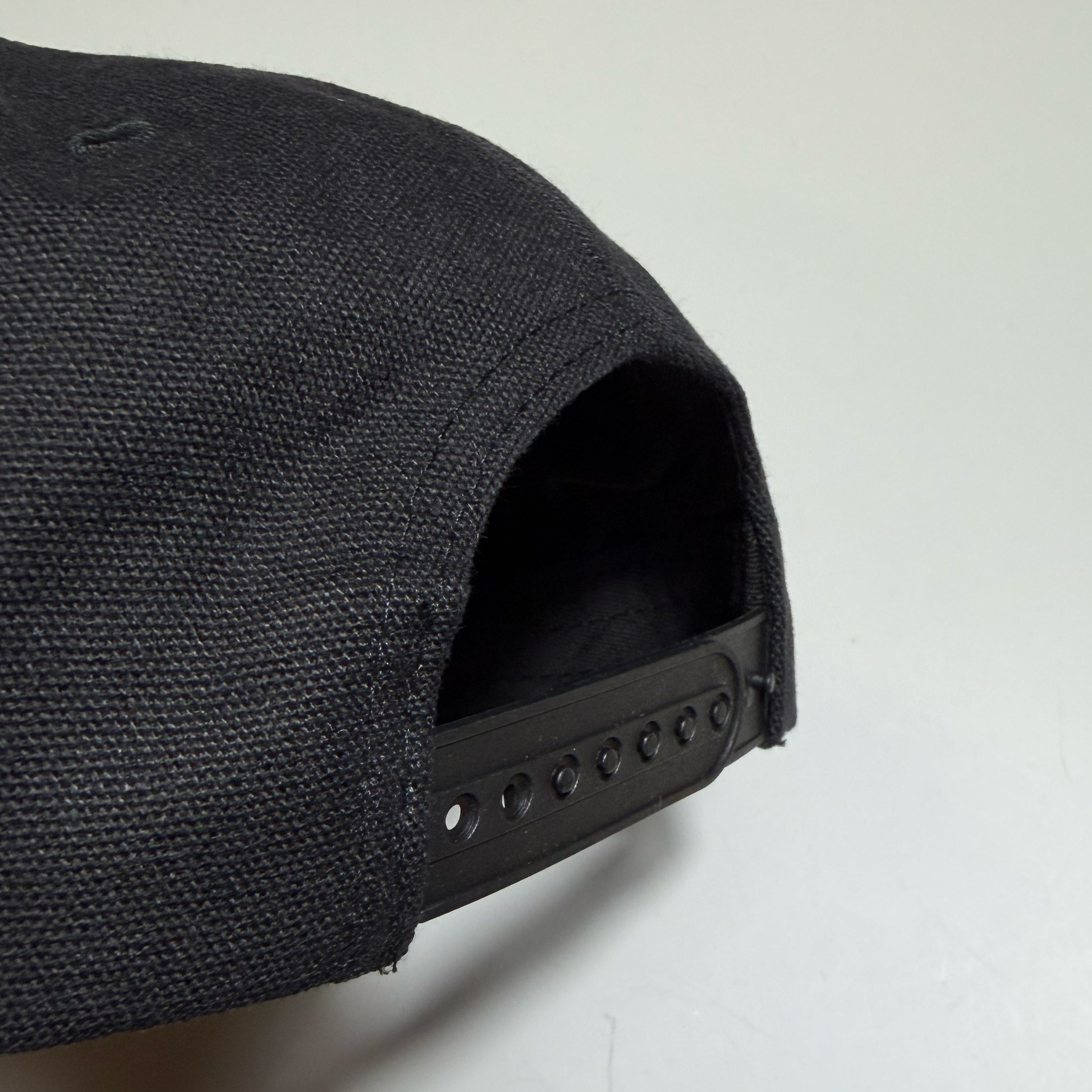 Close-up of the back adjustment area of a black textured baseball cap on a light gray background.