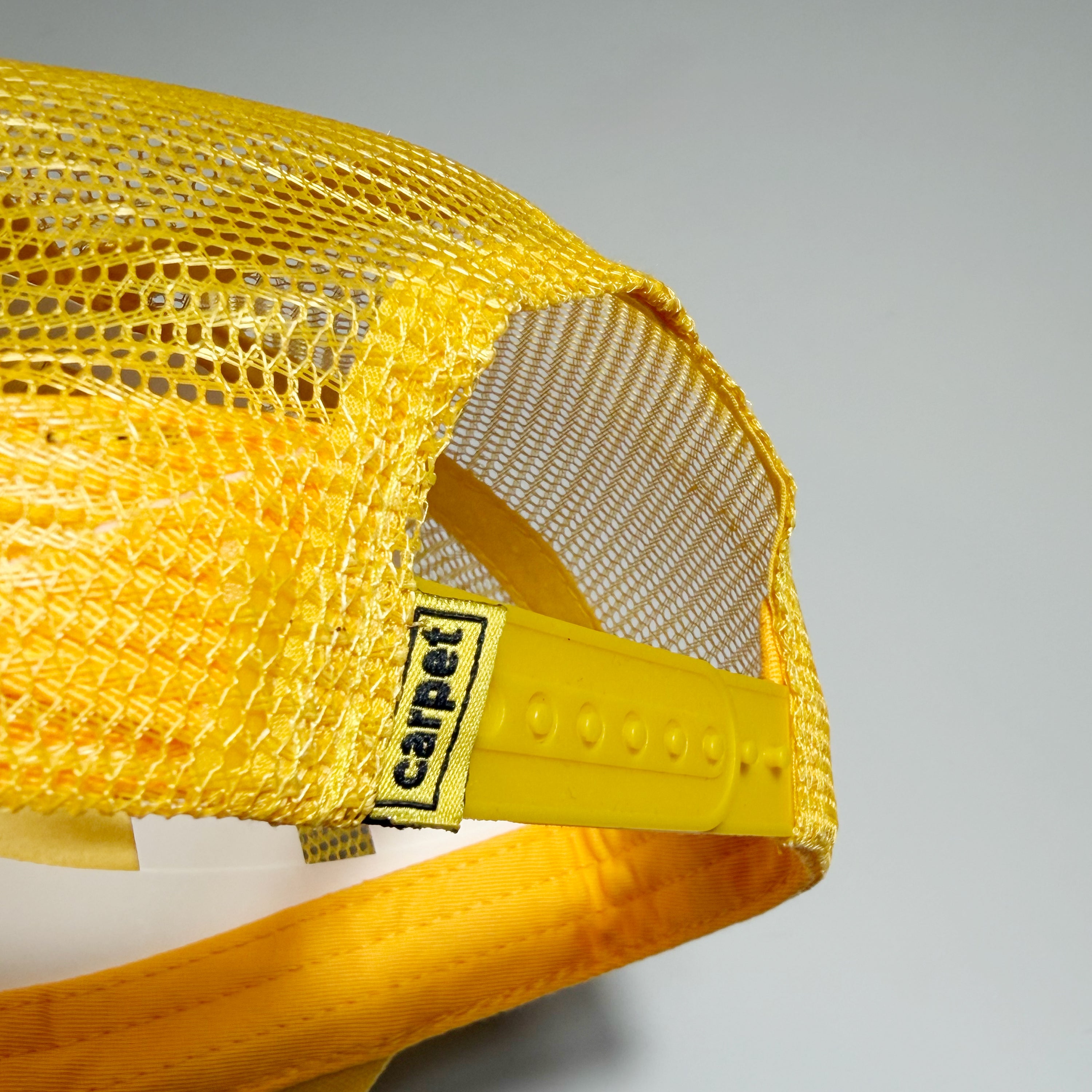 Close-up of a yellow mesh trucker cap showing the back mesh panels, yellow plastic back strap, and a black label with the brand name 'carpet'.