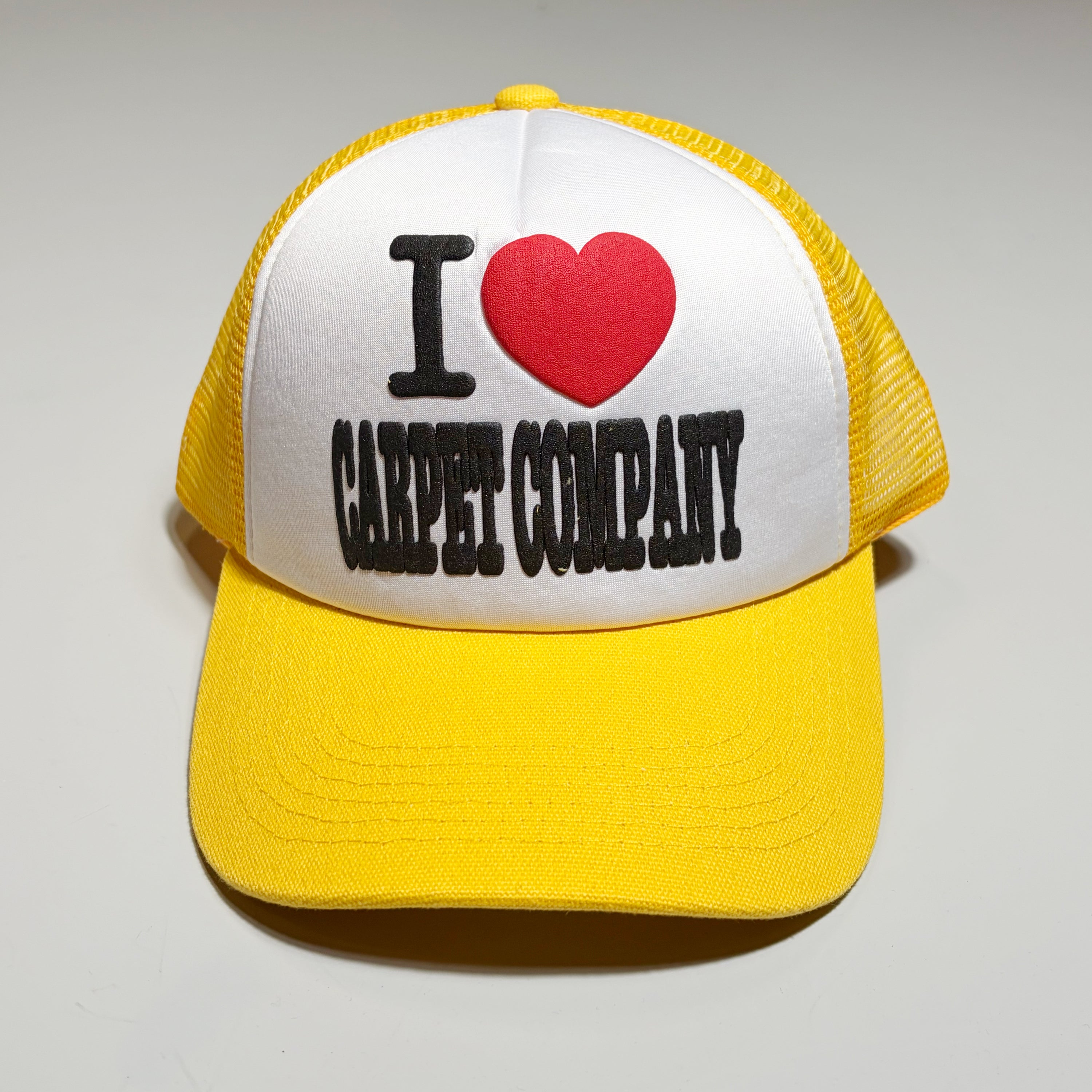 Front image of yellow/white trucker hat. I heart carpet company in puff printed ink on crown.
