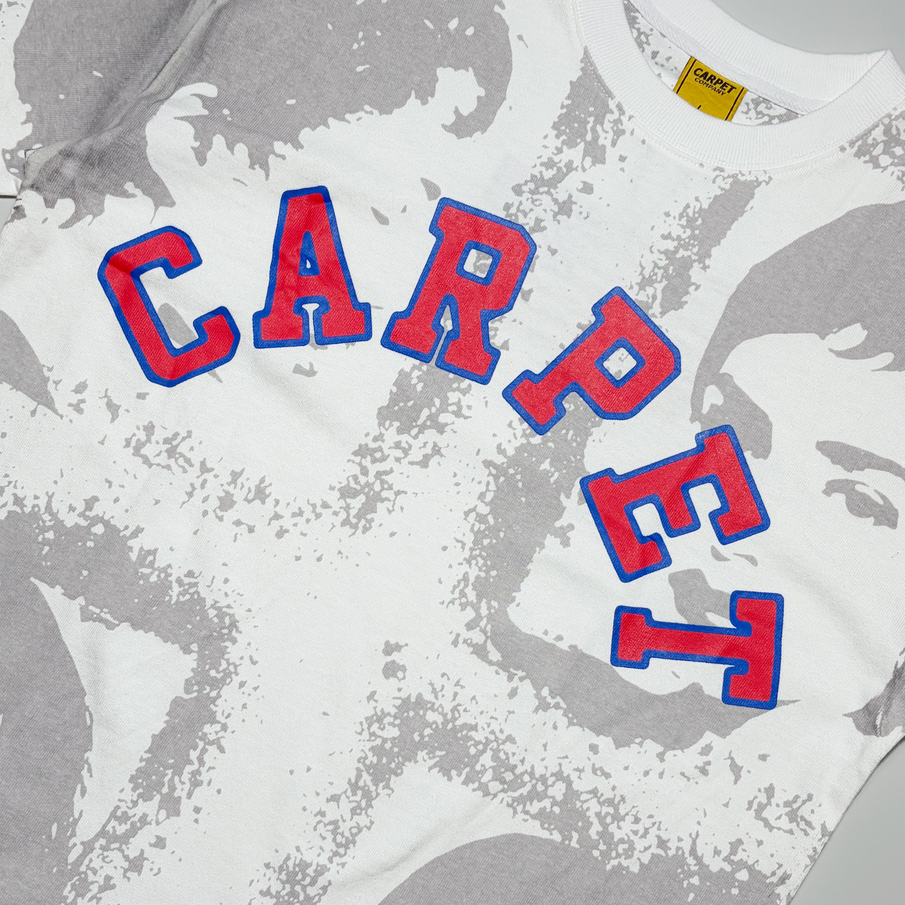 A close-up of a white t-shirt featuring a gray distressed graphic print and the word 'CARPET' arched across the chest in red varsity-style letters with a blue outline.