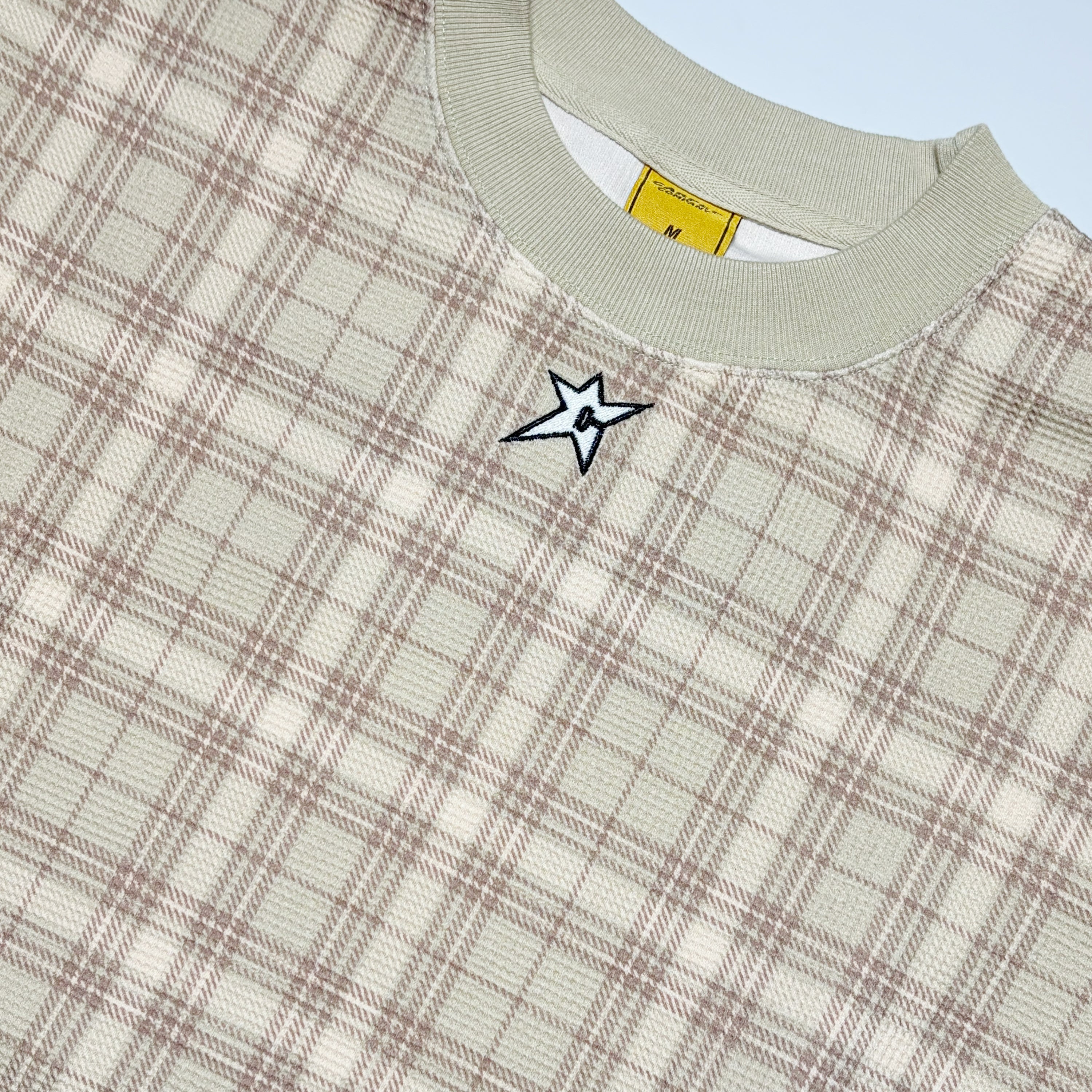 Close-up of a crewneck sweatshirt featuring a beige and brown plaid pattern, a light sage green ribbed collar, and a small embroidered white star with a black outline on the chest.