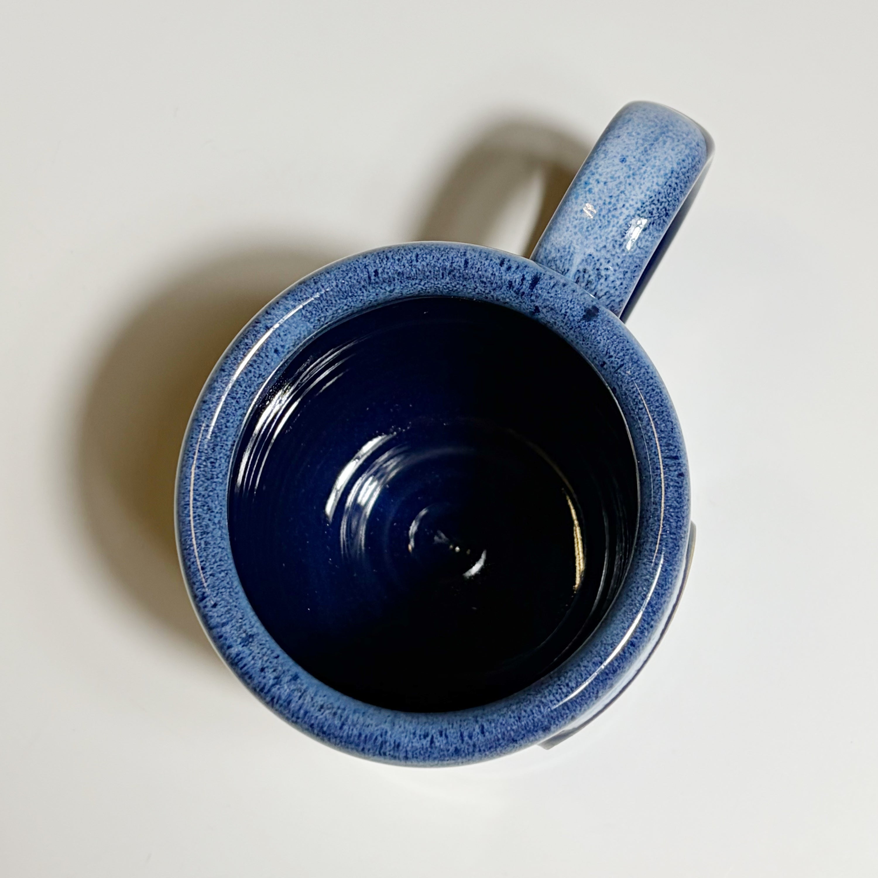 A ceramic mug with a speckled blue exterior and a glossy dark navy interior, photographed from a top-down angle on a light off-white background.