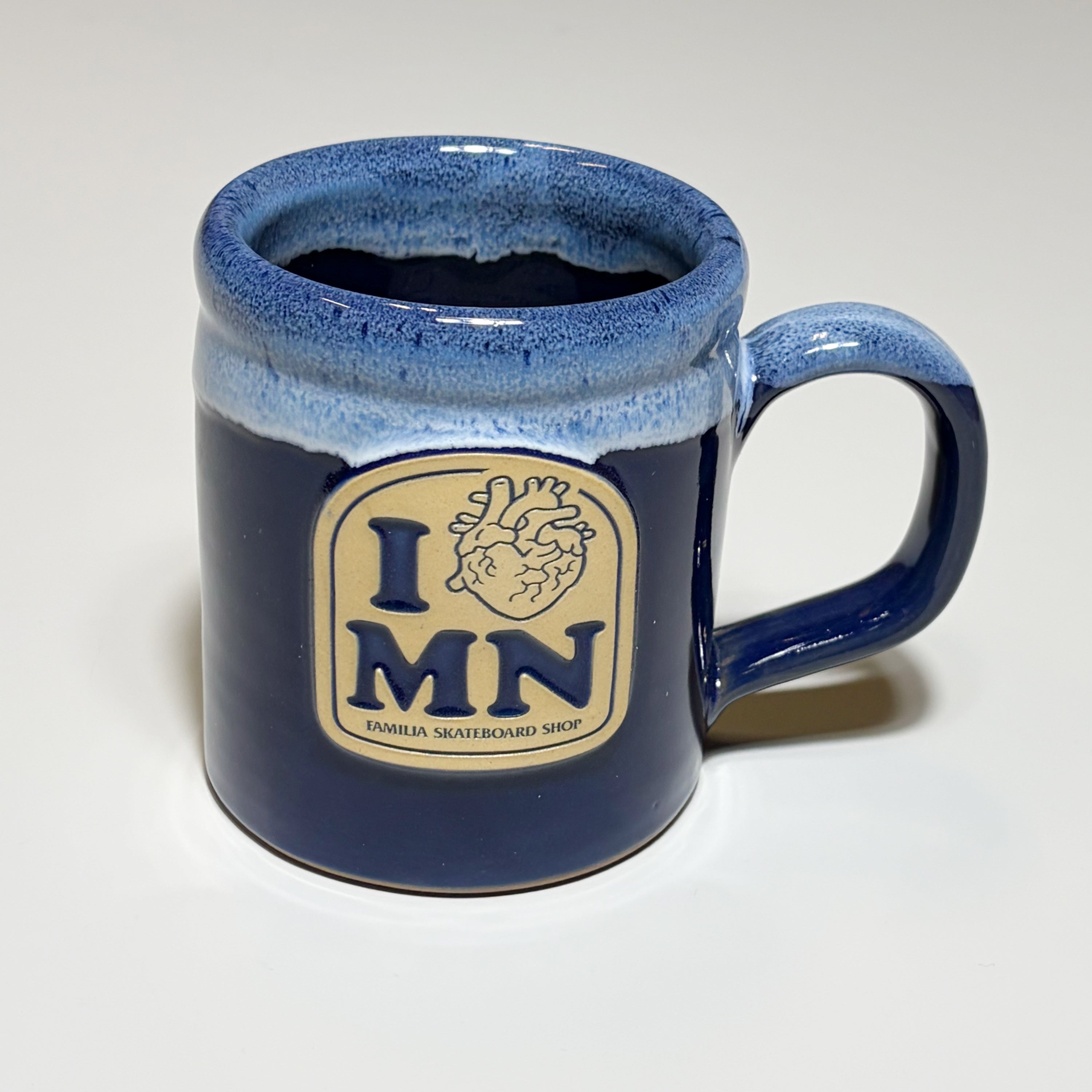 A dark blue glazed ceramic mug with a cream-colored 'I MN' logo patch on the front, featuring a heart graphic and the text 'FAMILIA SKATEBOARD SHOP'.