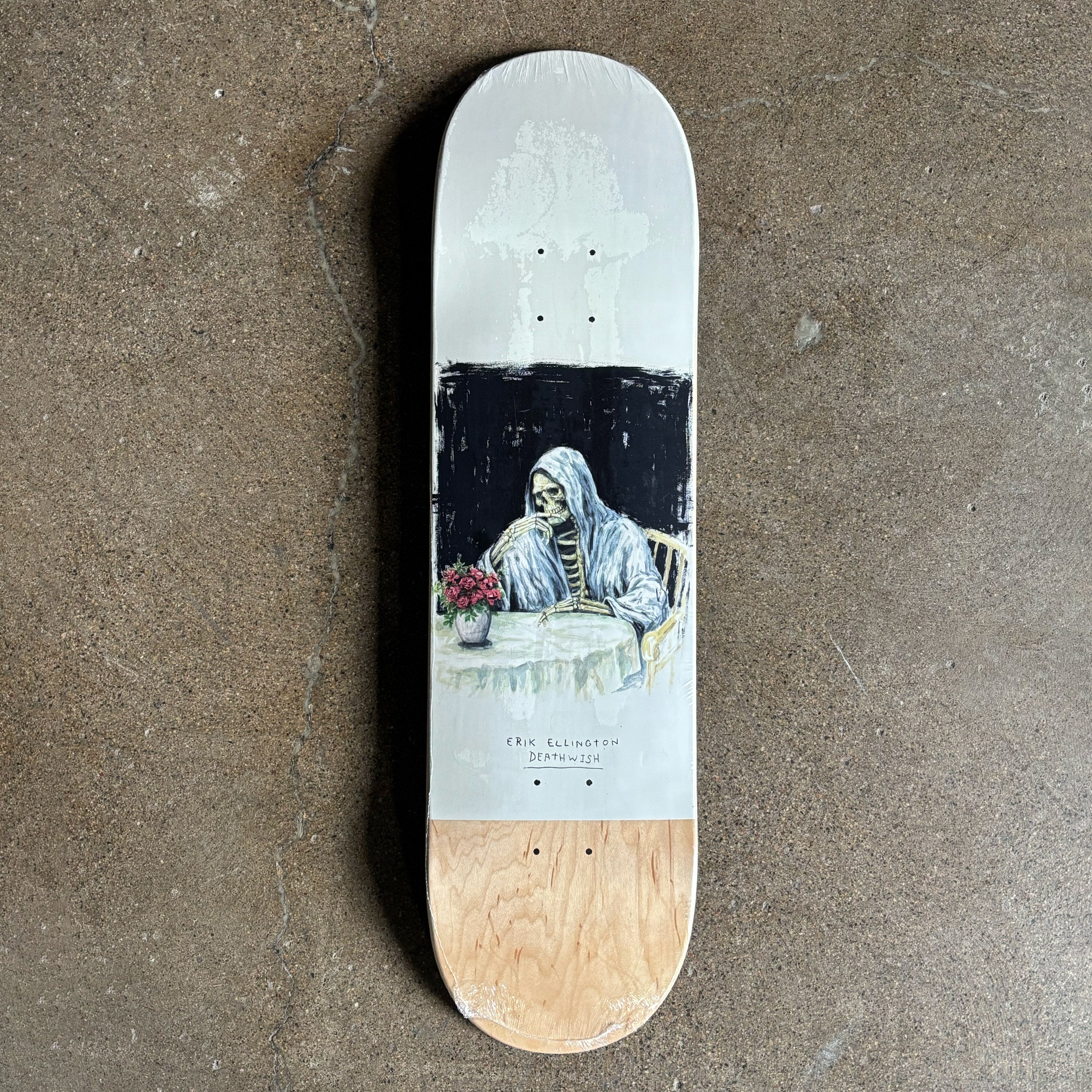 Skateboard deck with a skeleton at a table design on a concrete surface