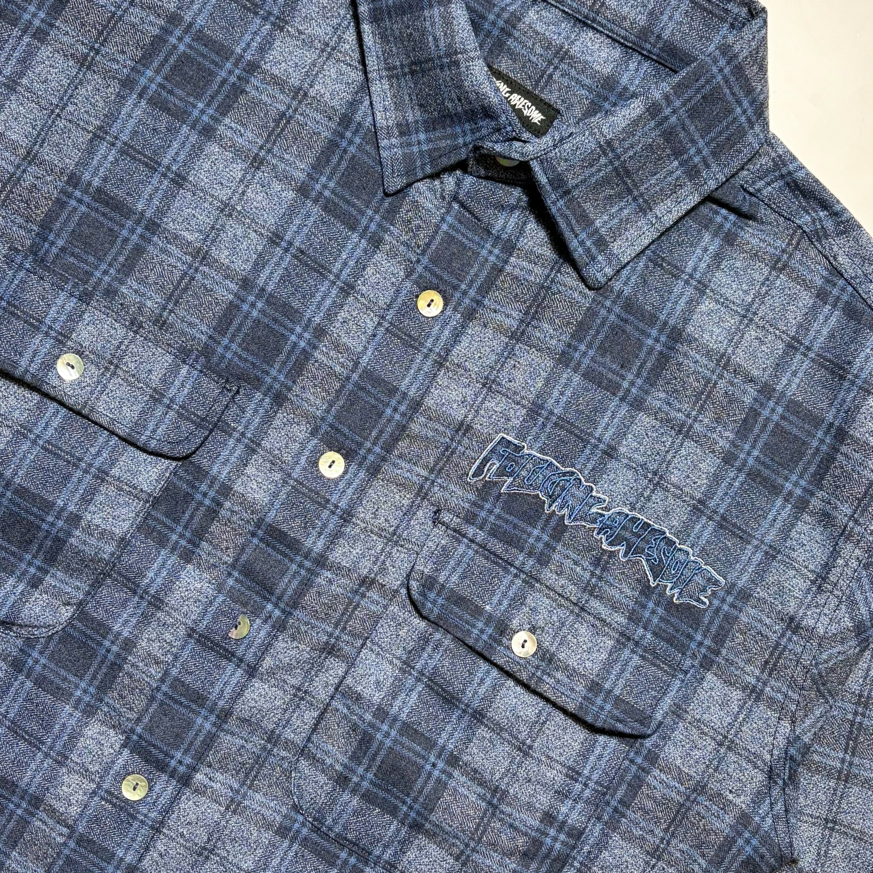 Close-up of a blue and gray plaid button-up shirt with a visible embroidered 'Fucking Awesome' logo on the chest pocket, laid flat on a light background.