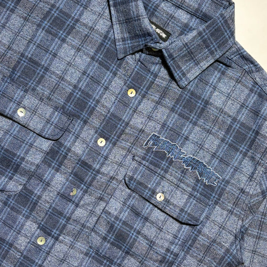 Close-up of a blue and gray plaid button-up shirt with a visible embroidered 'Fucking Awesome' logo on the chest pocket, laid flat on a light background.