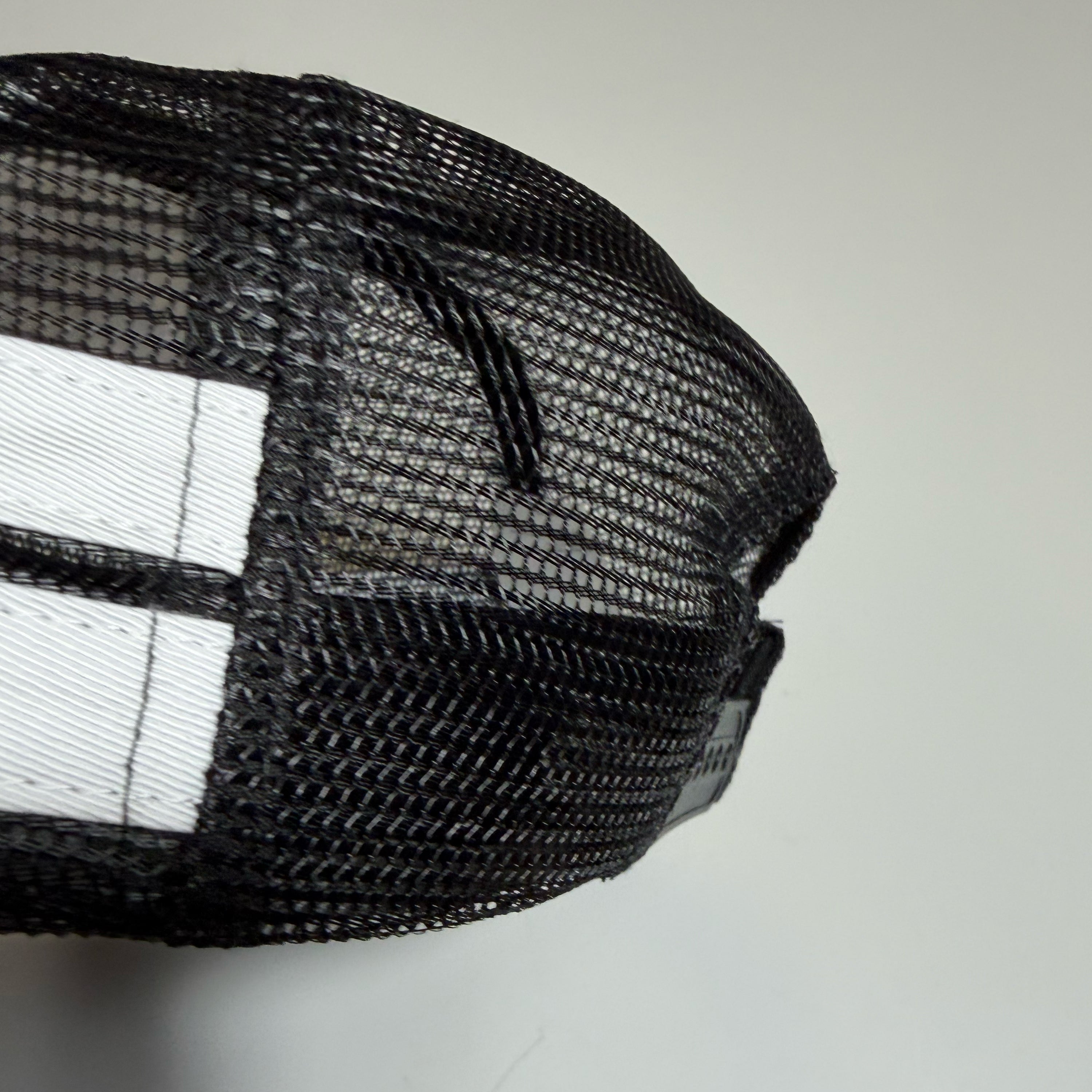 Close-up of a black mesh trucker hat showing the side and back panels with a white stripe detail on a light gray background.