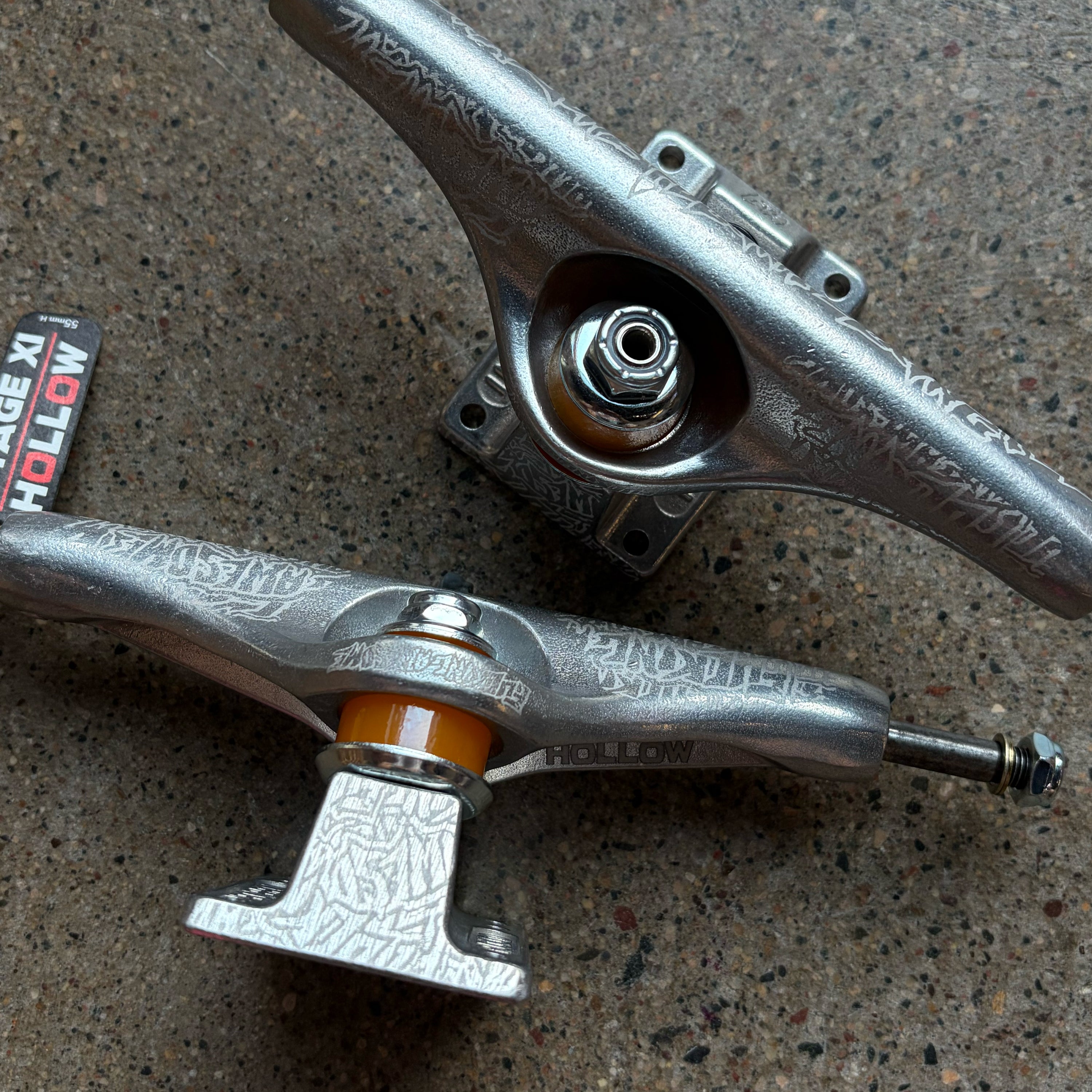 Two metallic silver skateboard trucks with engraved detailing and orange bushings, displayed on a textured concrete surface.