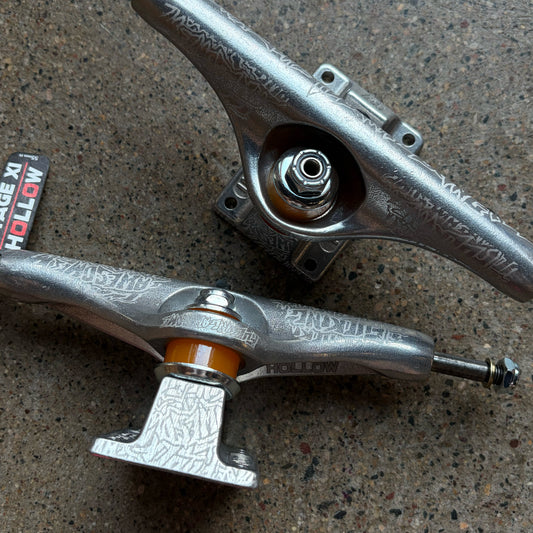 Two metallic silver skateboard trucks with engraved detailing and orange bushings, displayed on a textured concrete surface.