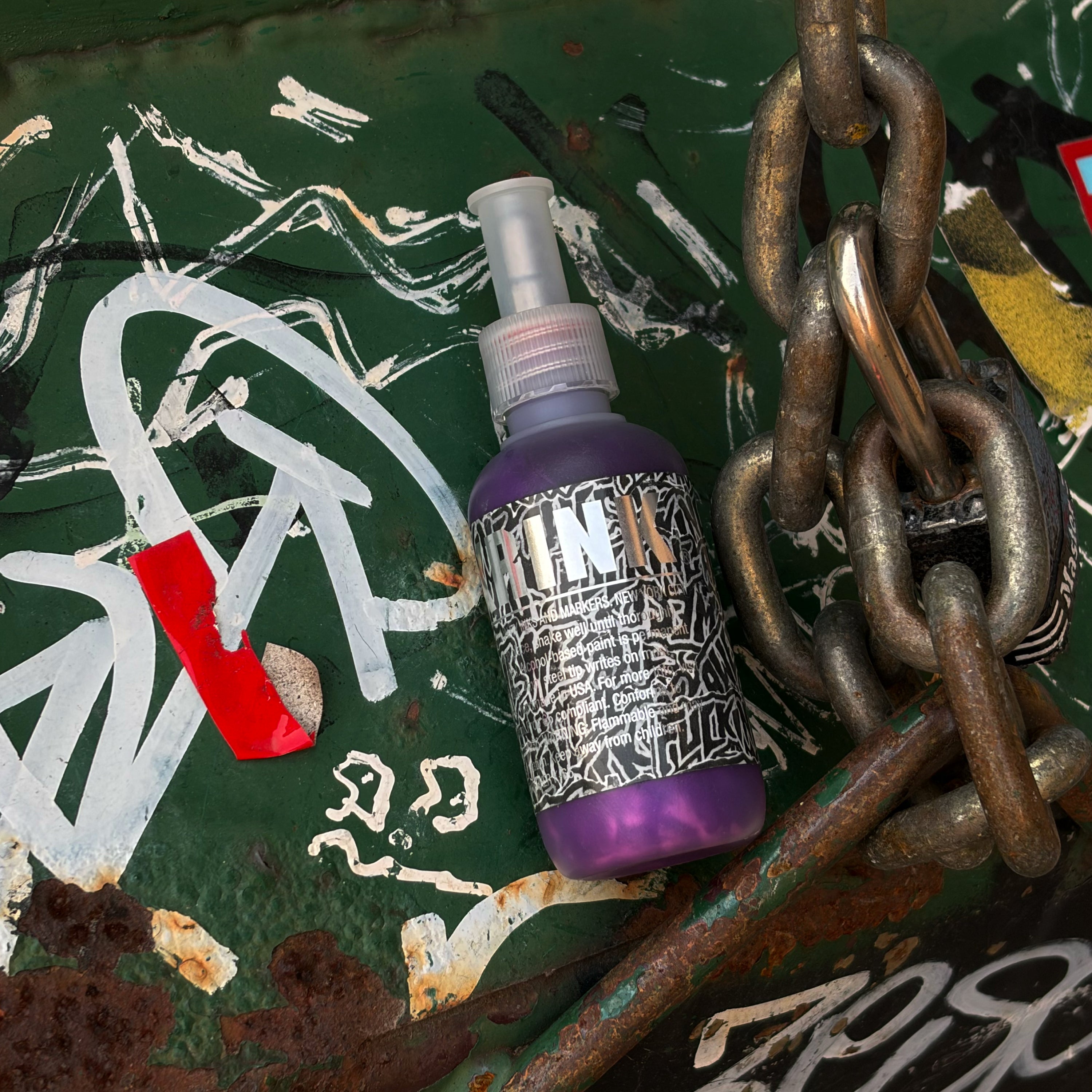 A purple spray bottle labeled 'Krink' with a black-and-white patterned label, placed on a distressed green graffiti surface with white paint marks, rust, and a large metal chain.