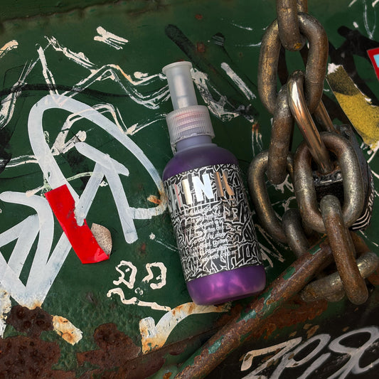 A purple spray bottle labeled 'Krink' with a black-and-white patterned label, placed on a distressed green graffiti surface with white paint marks, rust, and a large metal chain.