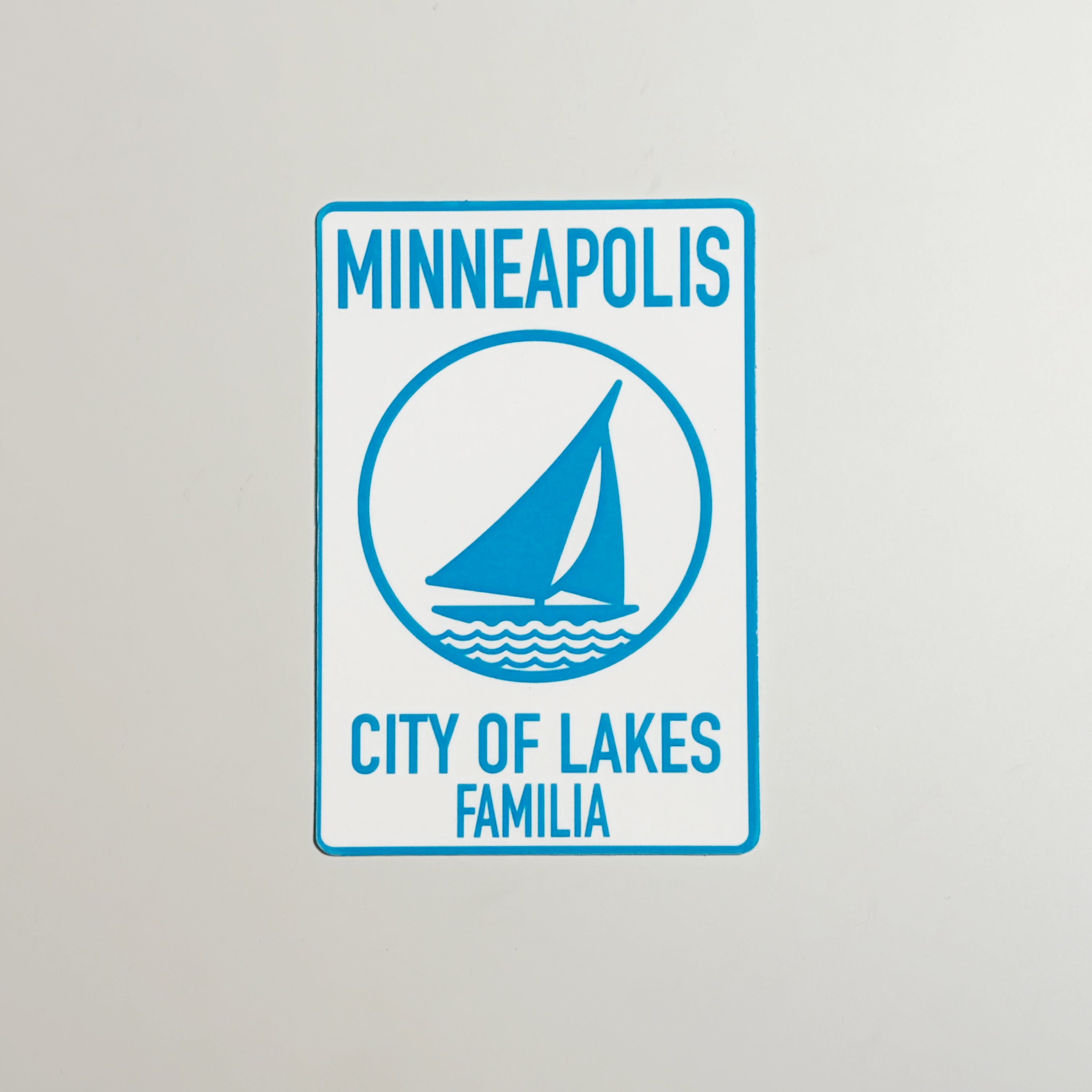 Sign with 'Minneapolis City of Lakes Familia' text and a sailboat icon on a gray background