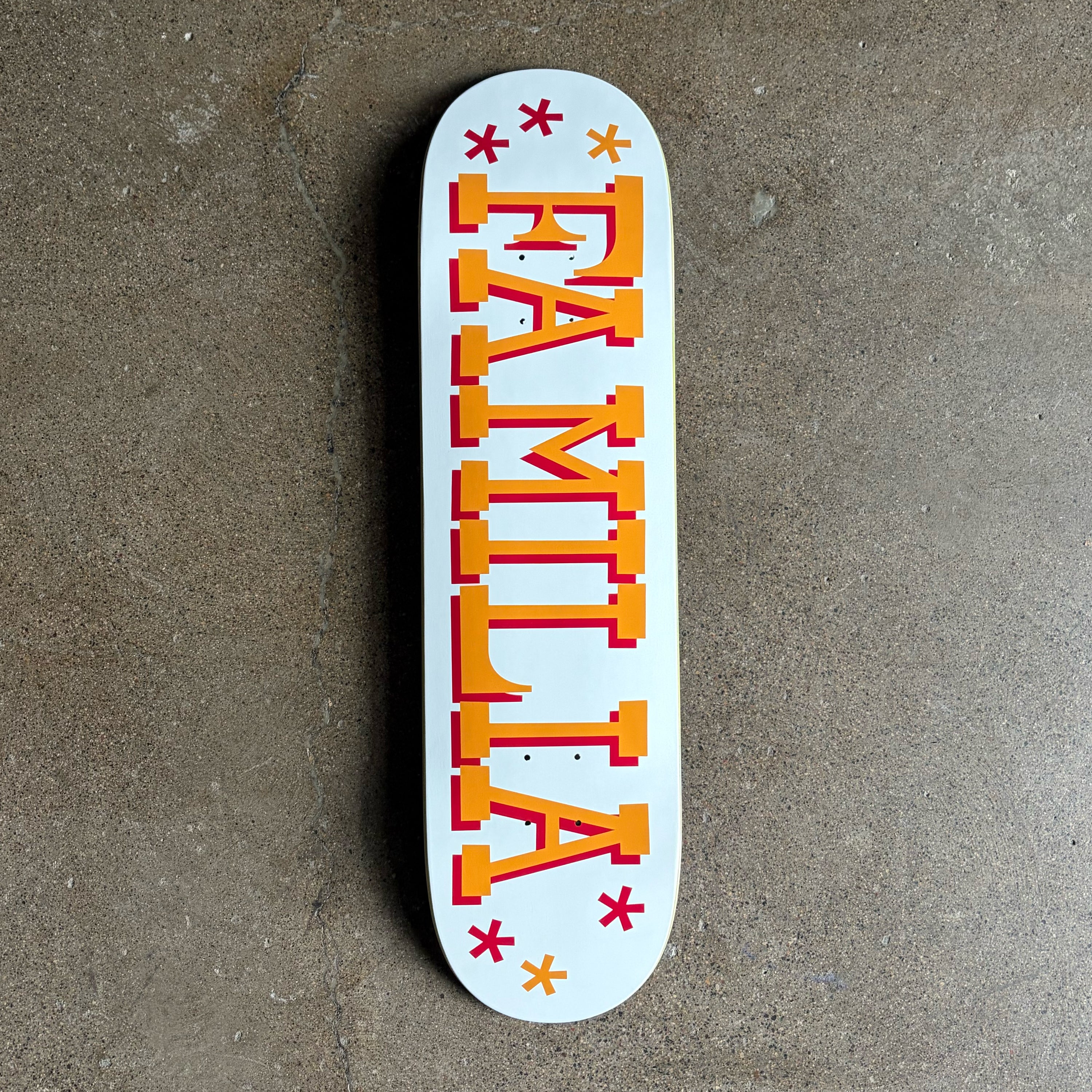 A white skateboard deck with the word 'FAMILY' printed vertically in bold orange letters with red drop shadows, accented by small red and orange stars, photographed from above on a gray concrete floor.