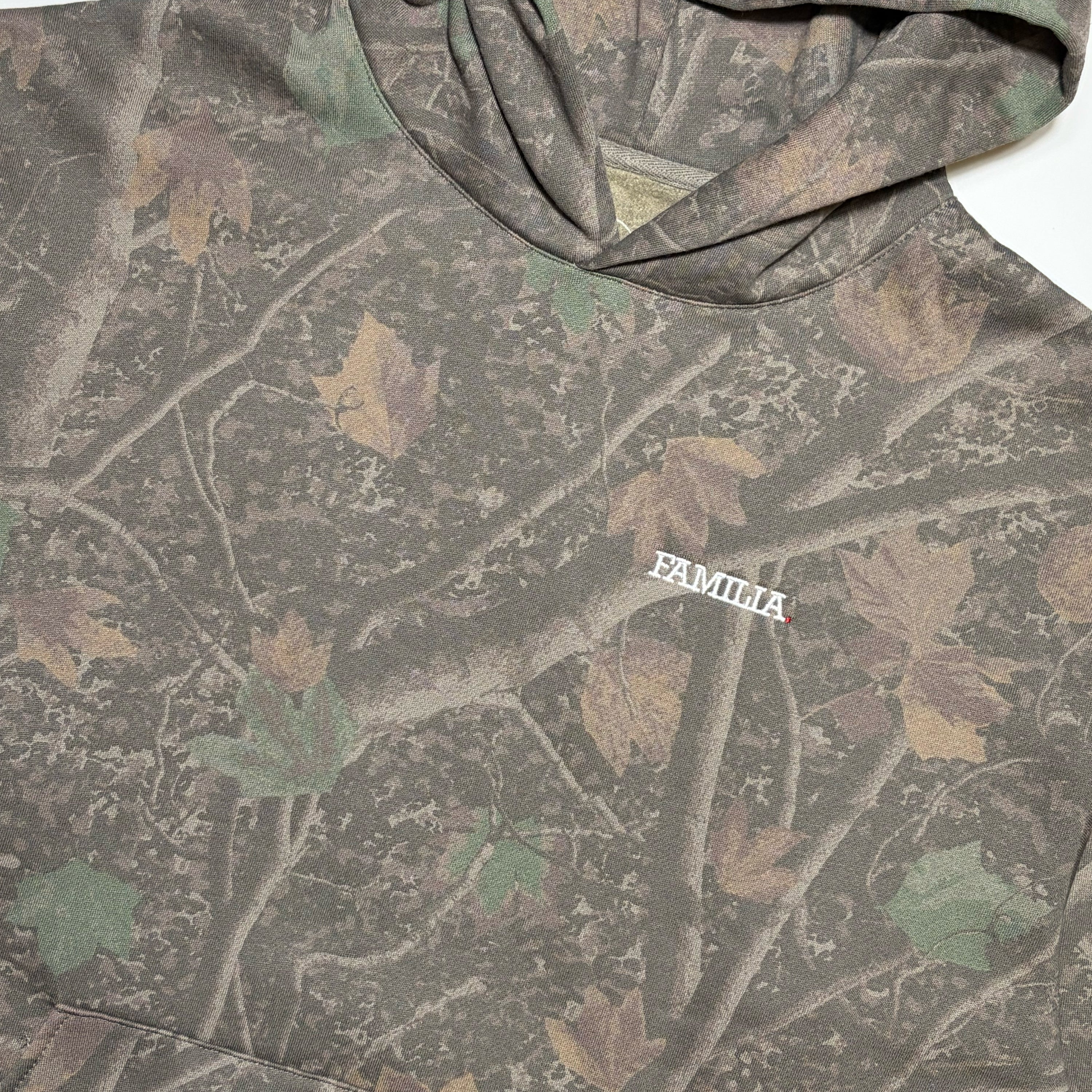 A close-up of a hooded sweatshirt featuring a woodland camouflage pattern with branches and leaves in muted green, brown, and tan. The word 'FAMILIA' is embroidered in white on the chest.