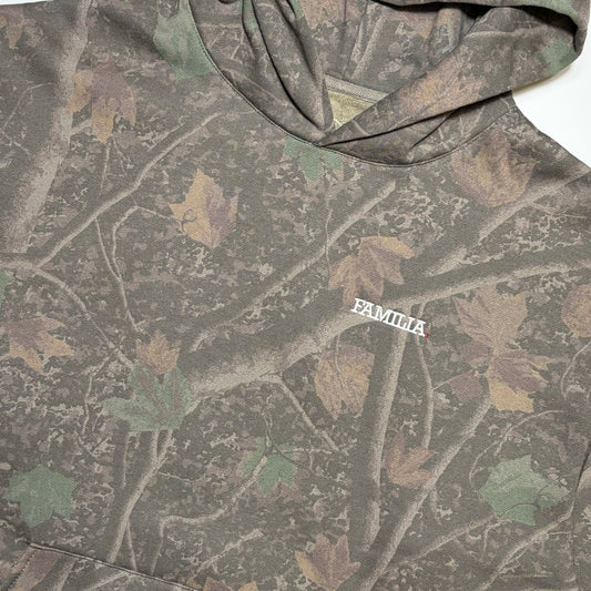 A close-up of a hooded sweatshirt featuring a woodland camouflage pattern with branches and leaves in muted green, brown, and tan. The word 'FAMILIA' is embroidered in white on the chest.