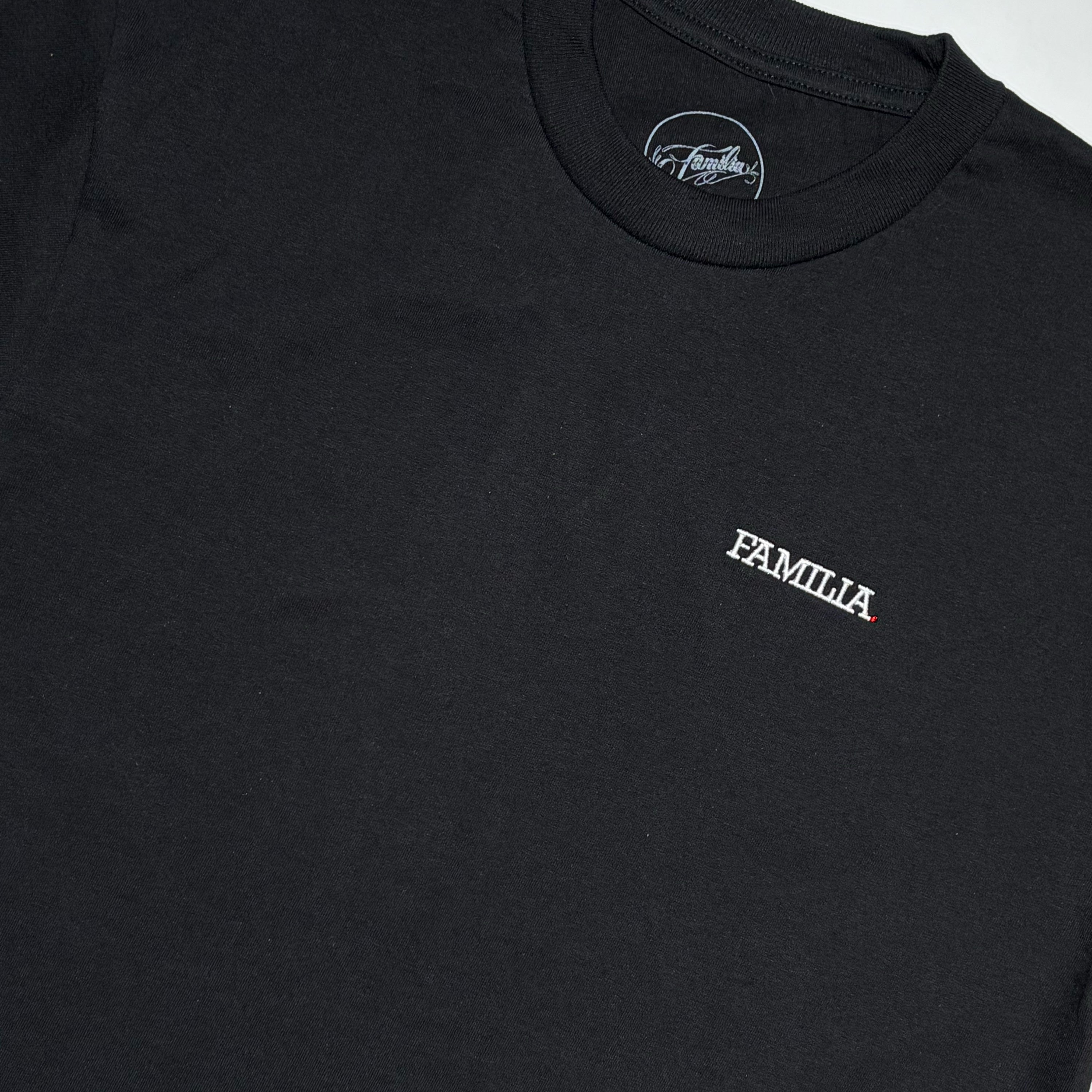 A close-up of a black t-shirt featuring the 'FAMILIA' logo embroidered in white on the left chest.