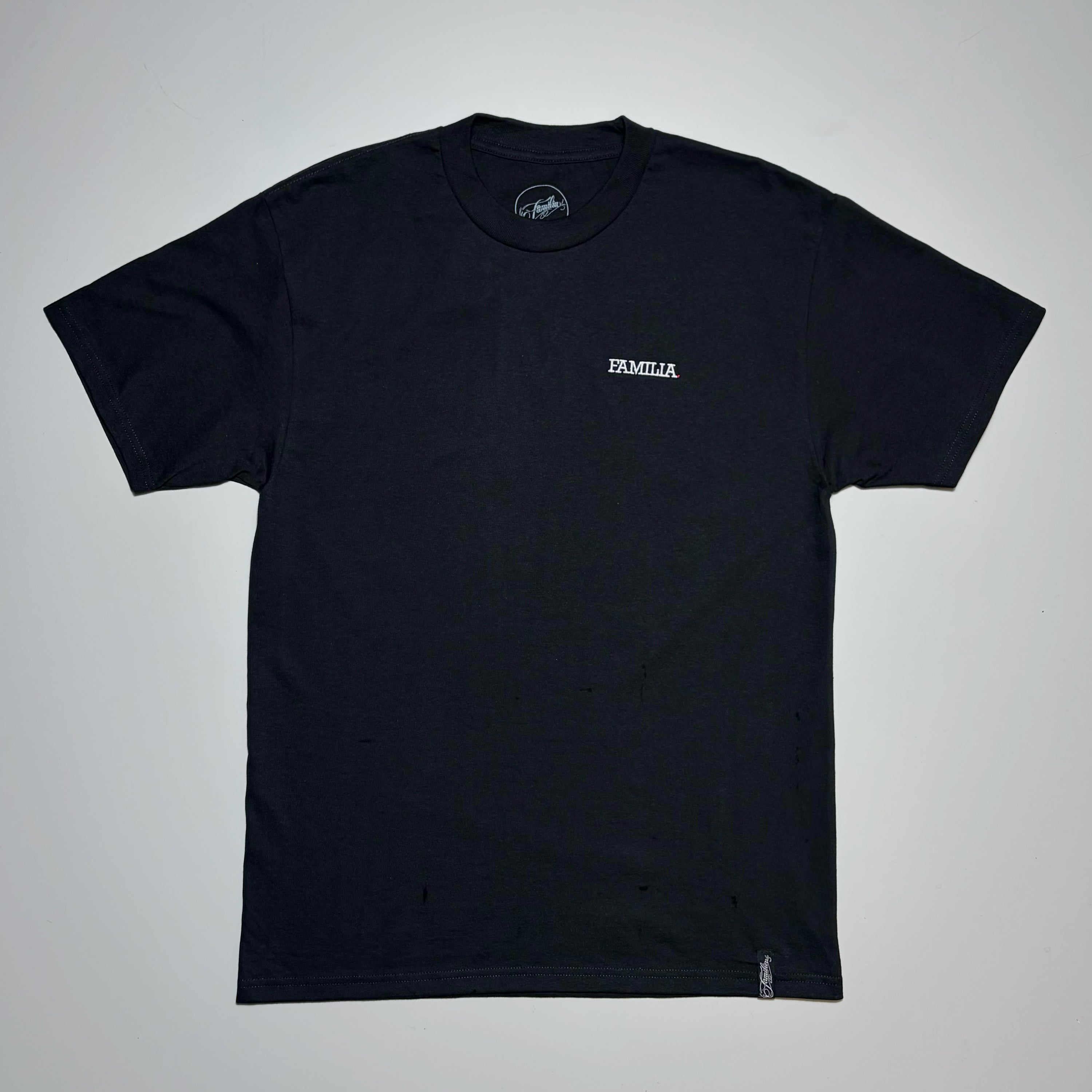 Full image of a black t-shirt with 'FAMILIA' embroidered in white on left chest. Laid flat on beige background
