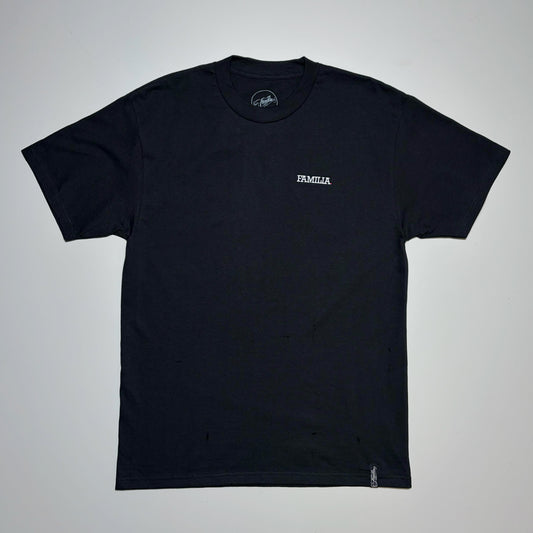 Full image of a black t-shirt with 'FAMILIA' embroidered in white on left chest. Laid flat on beige background